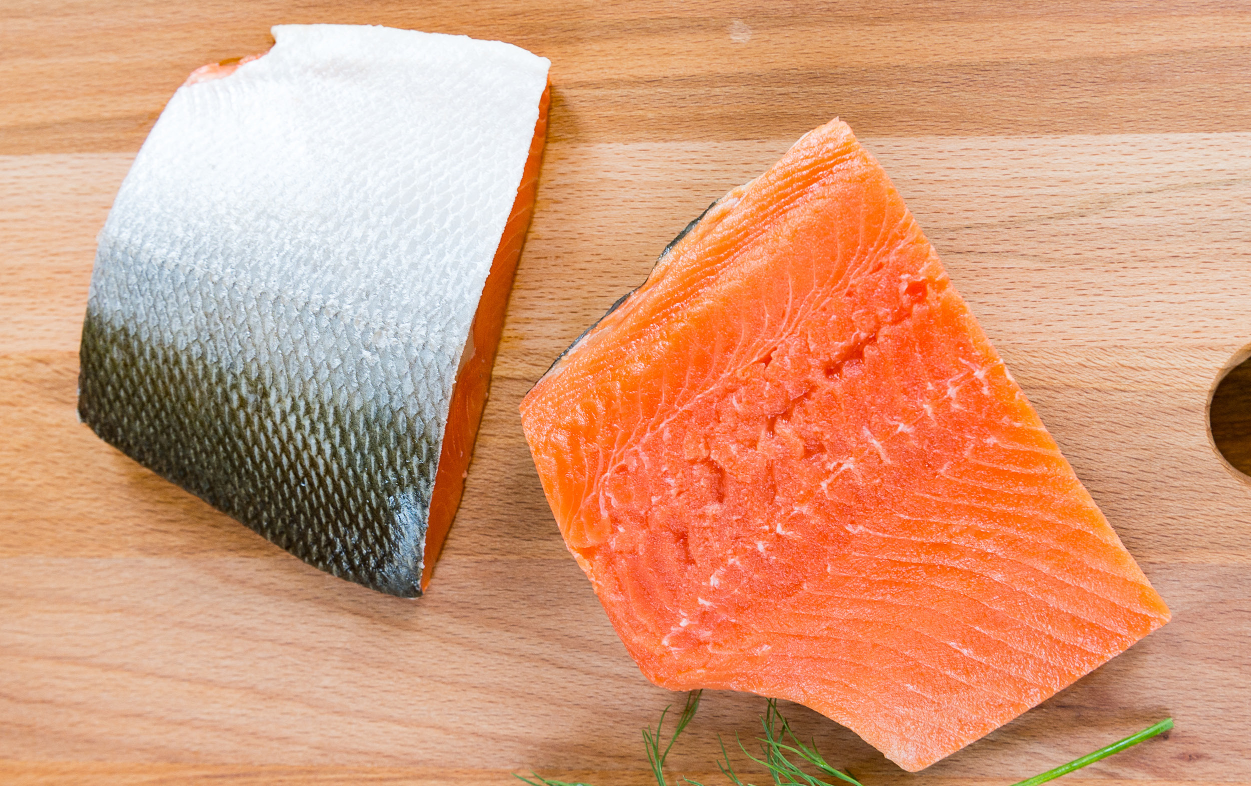 Two raw salmon fillets with skin on a wooden surface. One fillet shows the bright orange flesh, while the other highlights the silver skin. Some green herbs are partially visible in the corner.