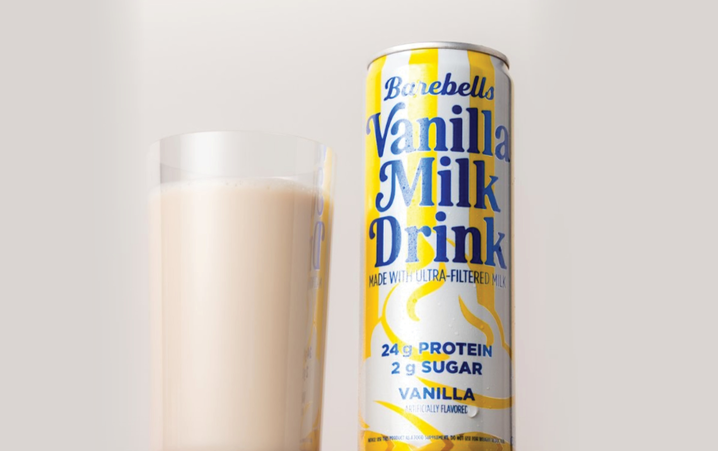 A can of Barebells Vanilla Milk Drink from the New Arrivals stands next to a glass filled with the light beige beverage. The can highlights 24g protein, 2g sugar, and vanilla flavor in blue and yellow text on a white background.