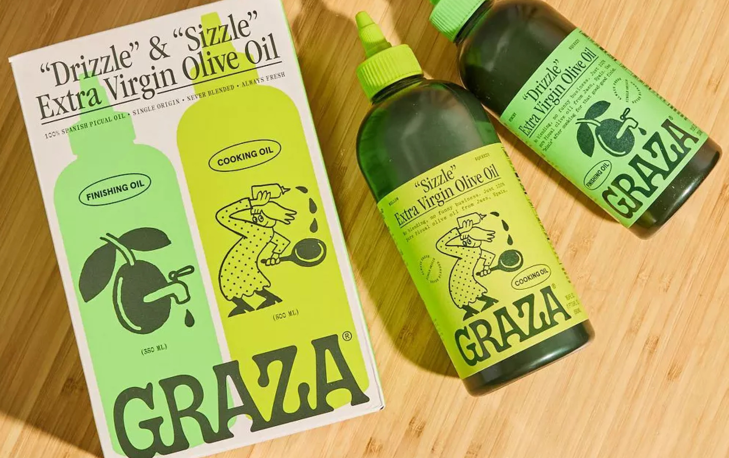 Two green bottles of Graza extra virgin olive oil, labeled Drizzle and Sizzle, sit next to their colorful box on a wooden surface. The playful packaging highlights these New Arrivals with bold text and lively illustrations.