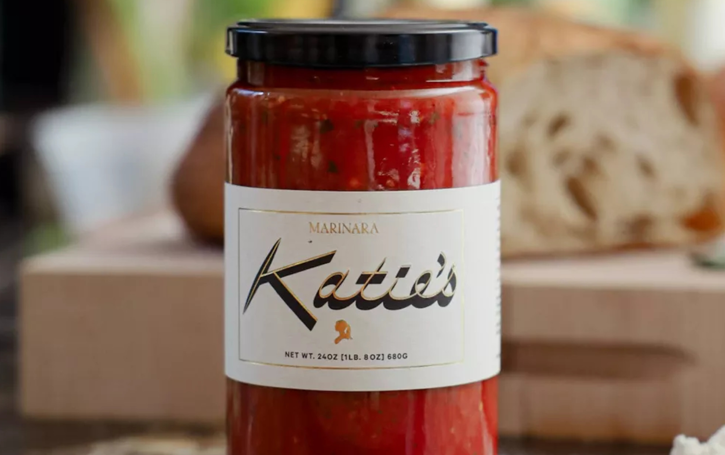 A jar of Katie’s marinara sauce with a black lid sits on a table, labeled “Katie’s” in script. Part of our New Arrivals, it’s paired perfectly with the fresh loaf of bread in the background. Weight and ingredient details are included.