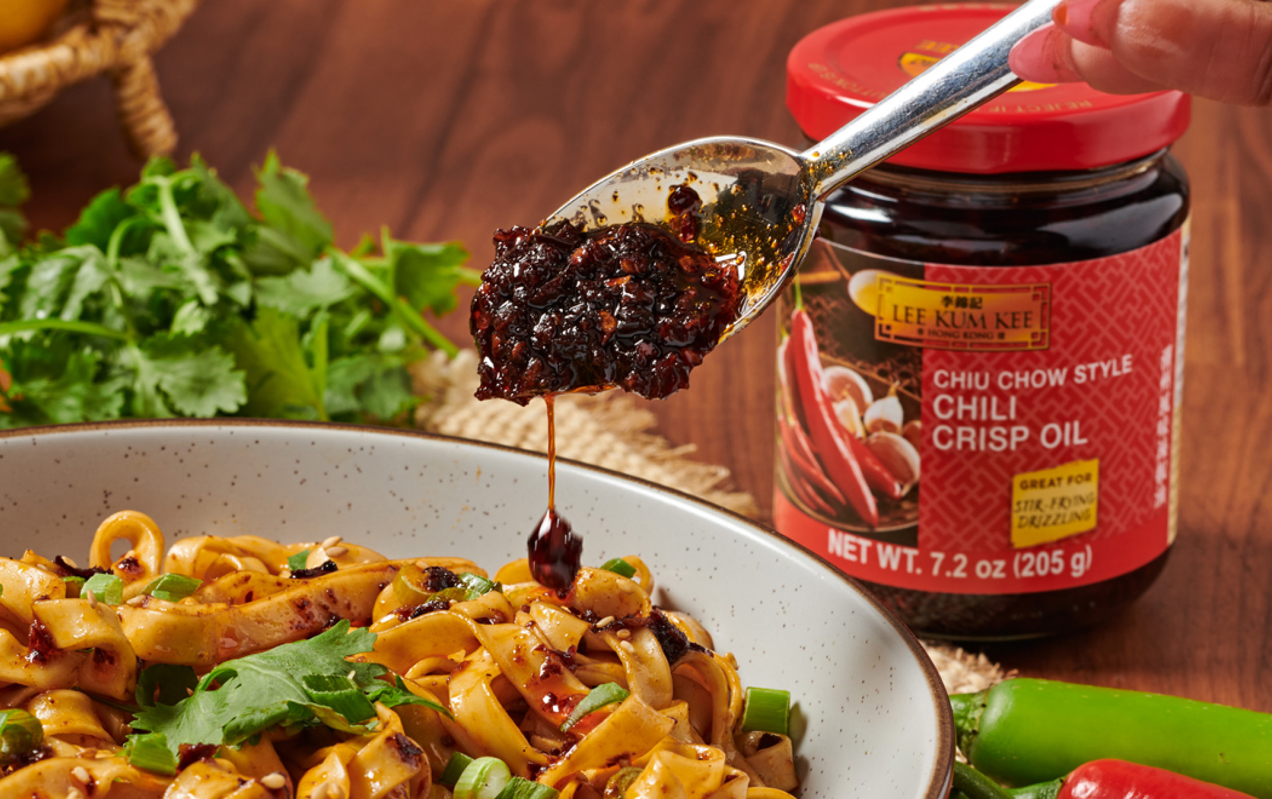 A spoonful of chili crisp oil is held above a bowl of noodles with vegetables and herbs. New Arrivals like Lee Kum Kee Chiu Chow Style Chili Crisp Oil are featured, with fresh cilantro and chili peppers nearby.