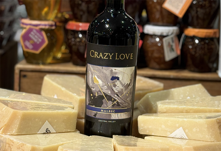 A bottle of Crazy Love Malbec wine is displayed surrounded by wedges of cheese, with jars of preserves in the background—perfect for anyone seeking Buy One Get One Wines to share and enjoy.