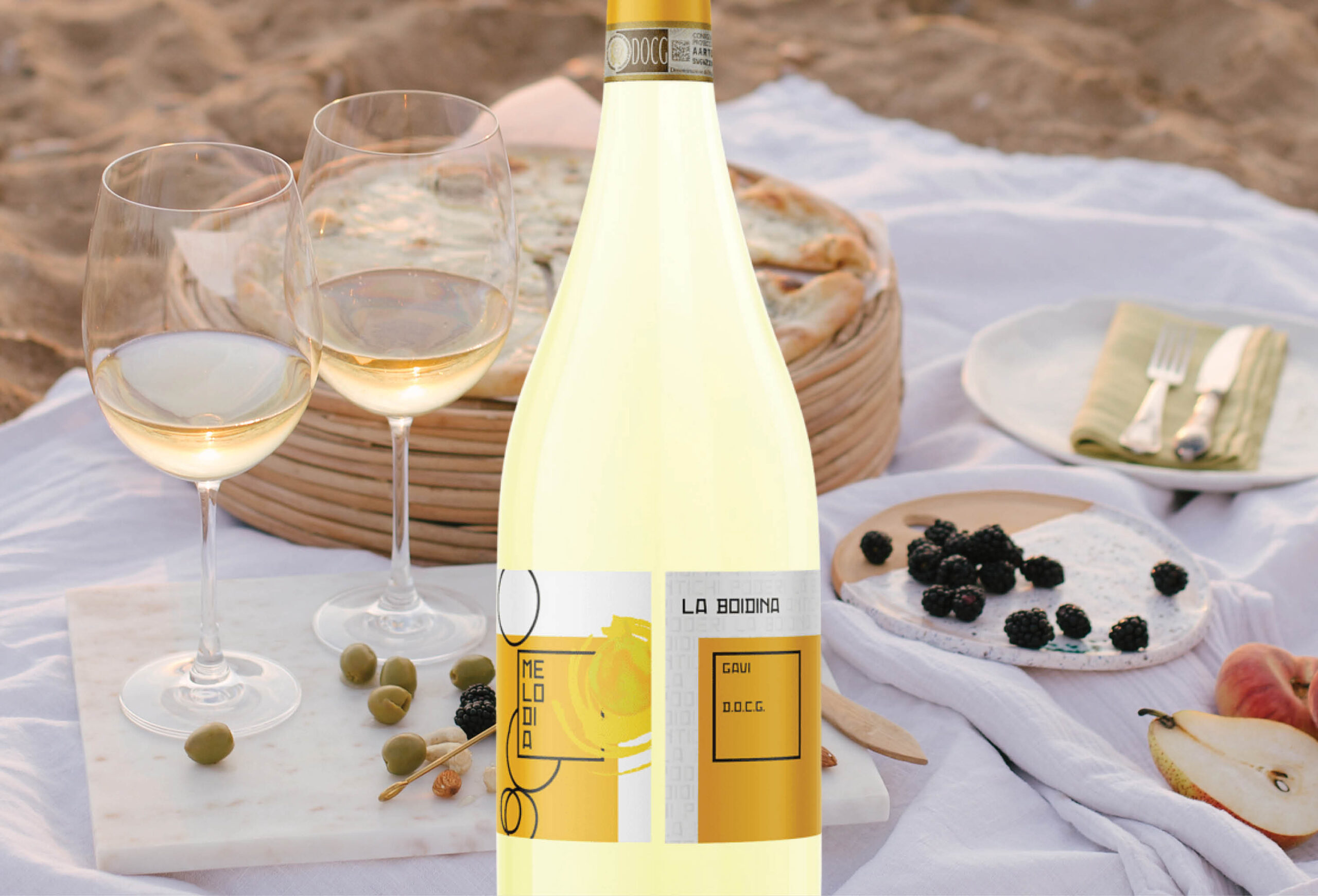 A bottle of white wine labeled La Bodina stands on a picnic setup with two glasses, a pie, cheese, blackberries, and green olives on a white cloth at the beach—perfect for enjoying with a special Buy One Get One Free Wine Offer.