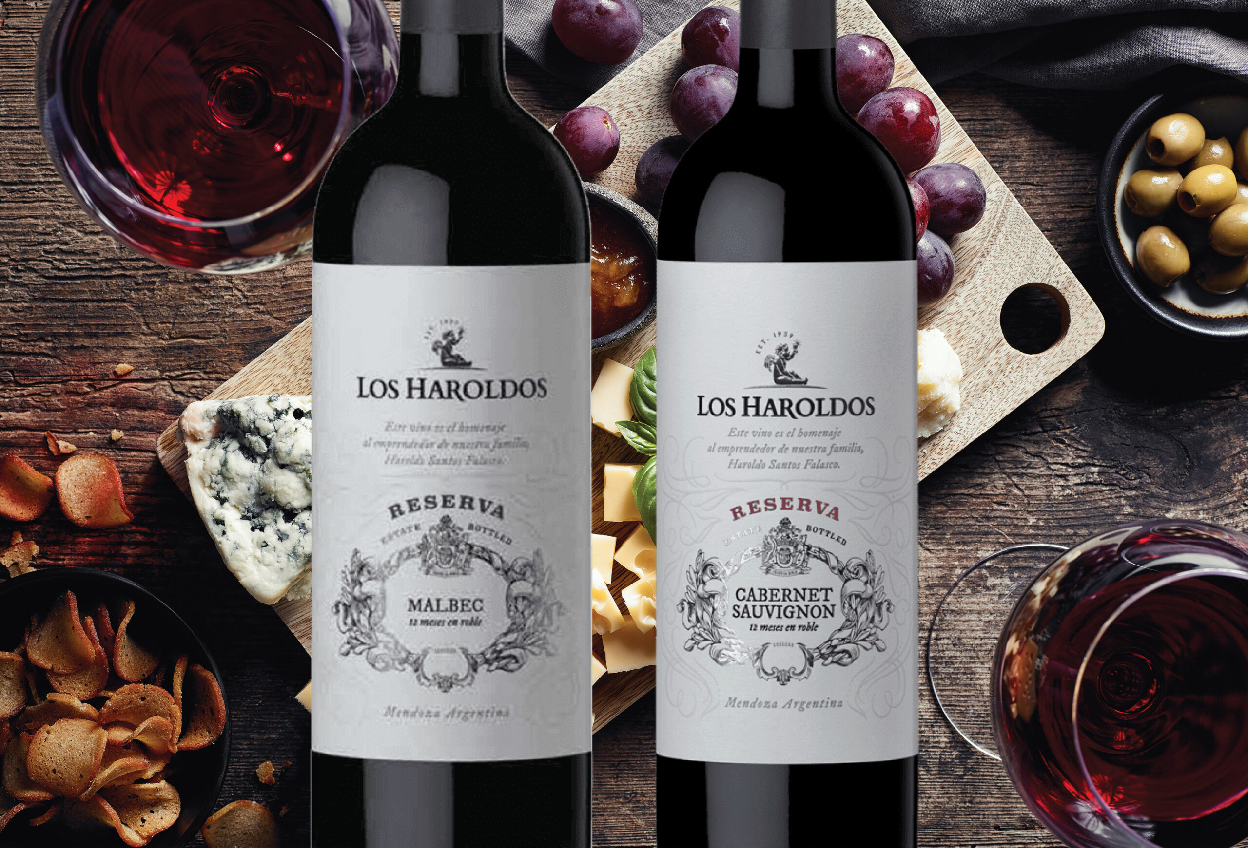 Two bottles of Los Haroldos Reserva wine, Malbec and Cabernet Sauvignon, are placed on a table with red wine glasses, assorted cheeses, grapes, olives, and crackers in the background.
