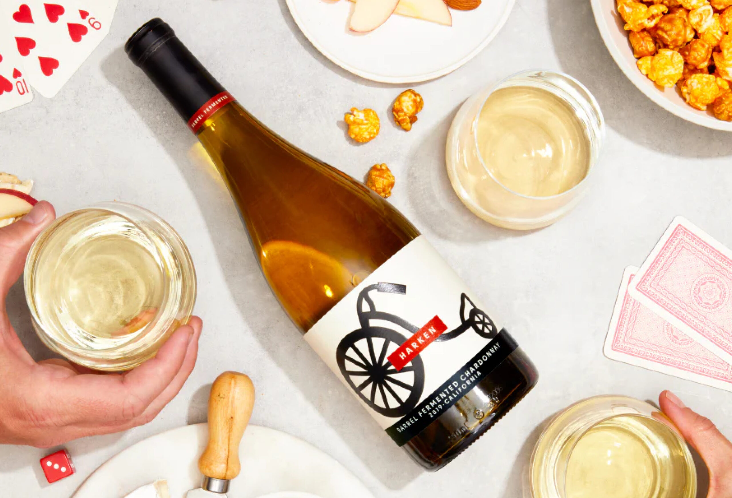 A bottle of Maker Chardonnay lies on a table surrounded by glasses of white wine, caramel popcorn, apple slices, a cheese knife, and playing cards, with hands holding two of the wine glasses.