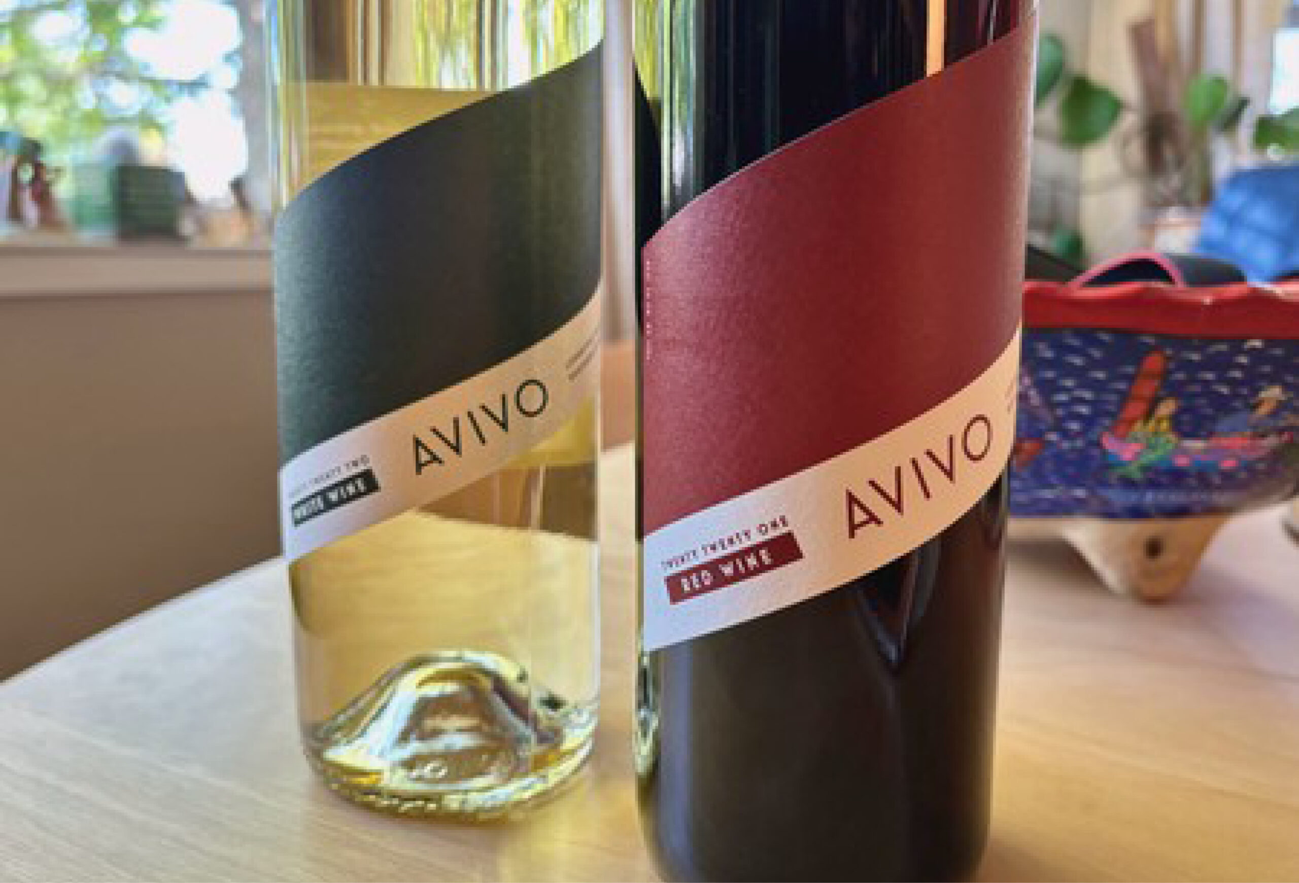 Two bottles of AVIVO wine on a table, one with a green label for white wine and the other with a red label for red wine. A colorful bowl and plants are in the background—perfect for enjoying exclusive Wine Deals.