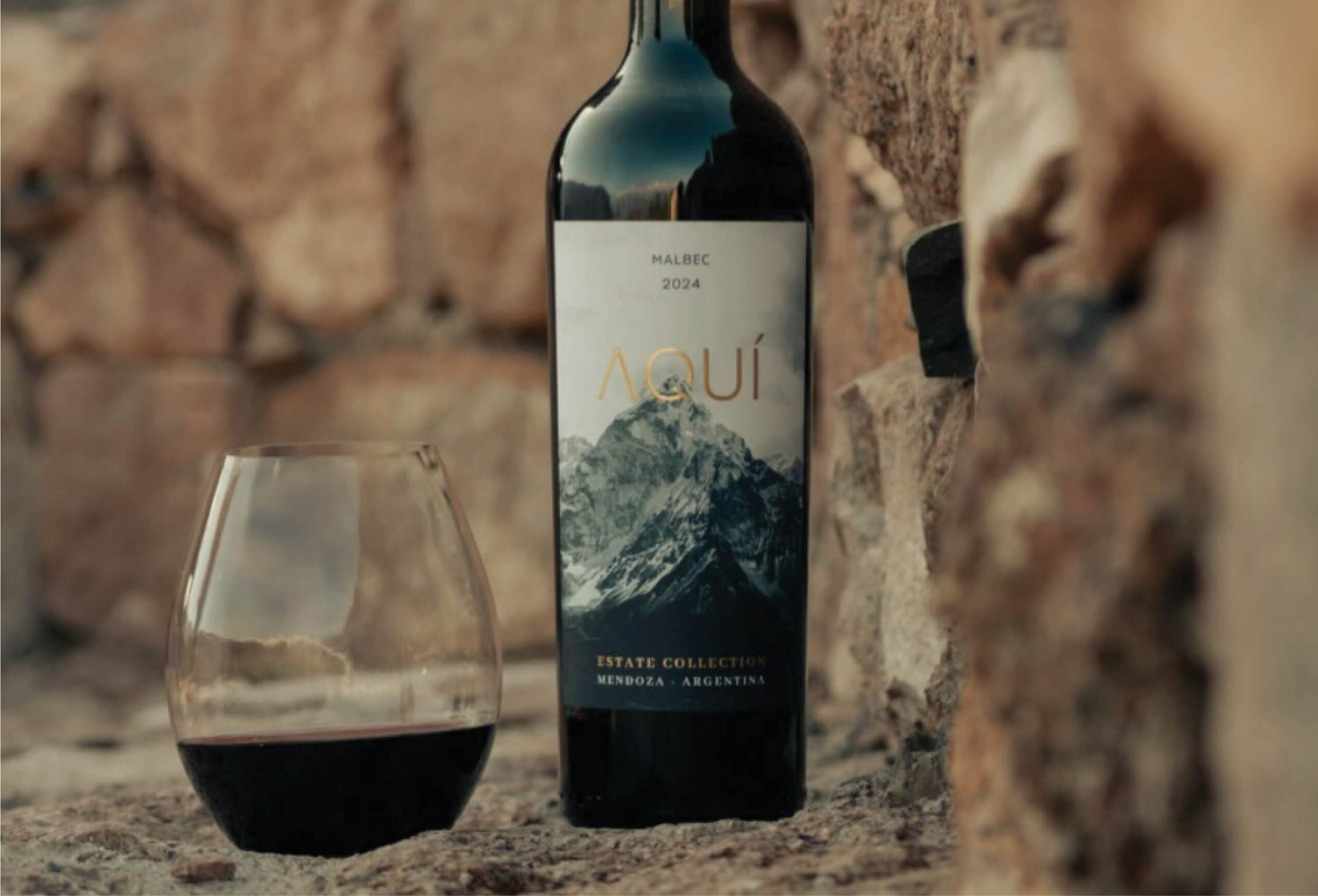 A bottle of Aquí Malbec 2024 wine and a glass of red rest on a stone surface, with rocky walls behind. The label features mountains and notes “Estate Collection, Mendoza, Argentina.” Discover exclusive Wine Deals when you buy today.