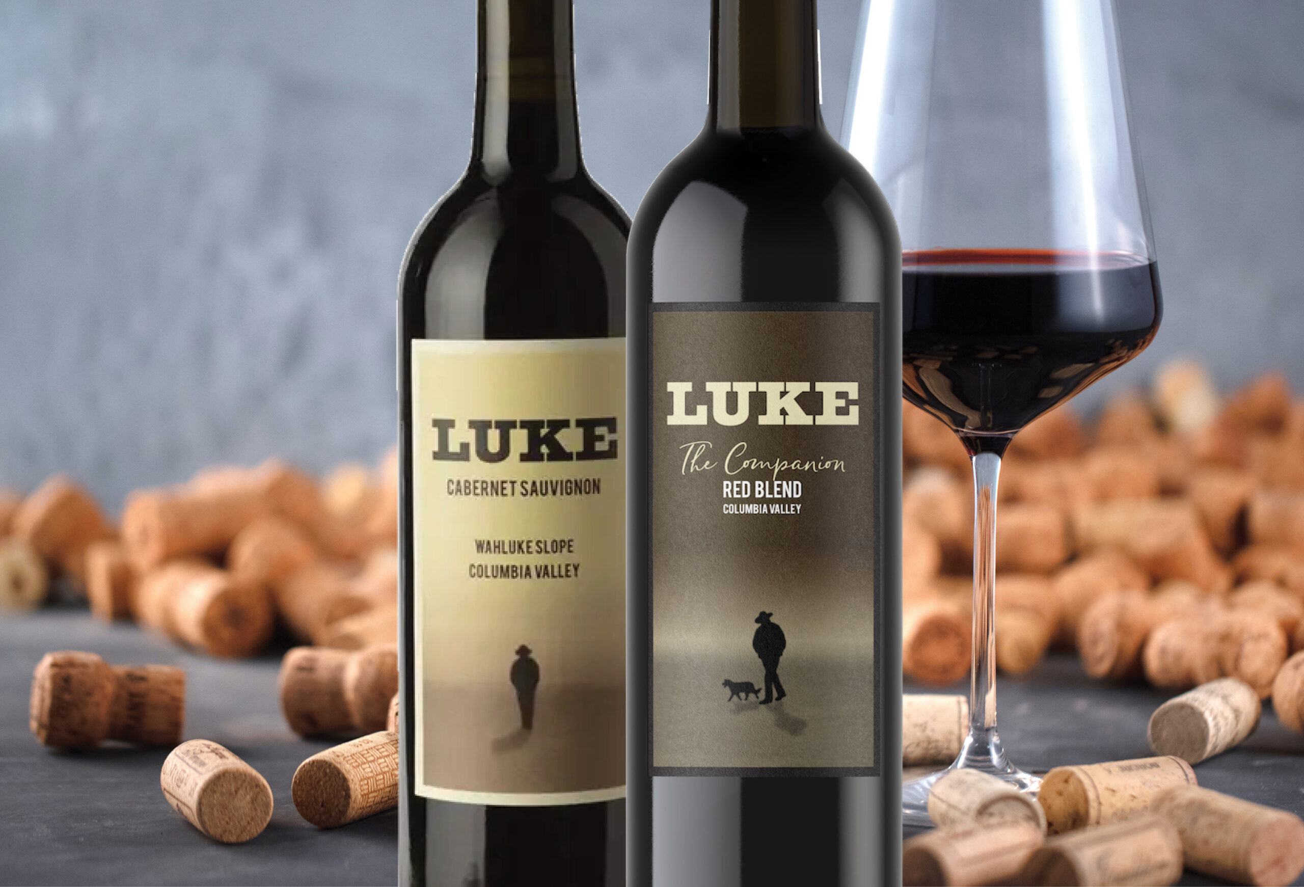 Two bottles of LUKE wine, one Cabernet Sauvignon and one Red Blend, stand side by side with a glass of red wine and scattered corks—discover exclusive wine deals on these beautifully labeled bottles featuring a silhouette of a person and a dog.