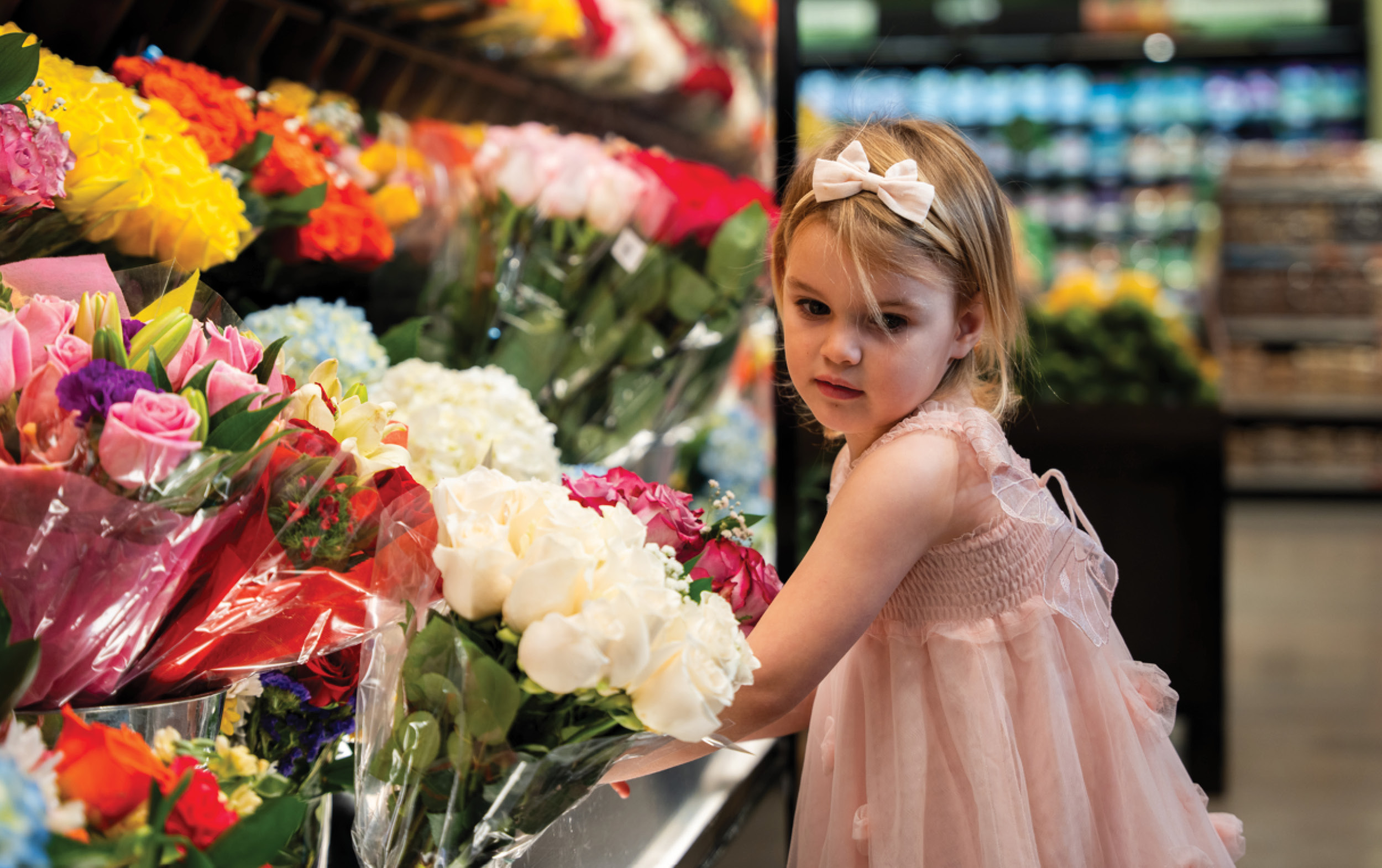 A young girl in a pink dress with a matching bow is standing in the grocery store flower section, joining other Little Shoppers as she admires bouquets of colorful flowers, including roses and hydrangeas, displayed on shelves.
