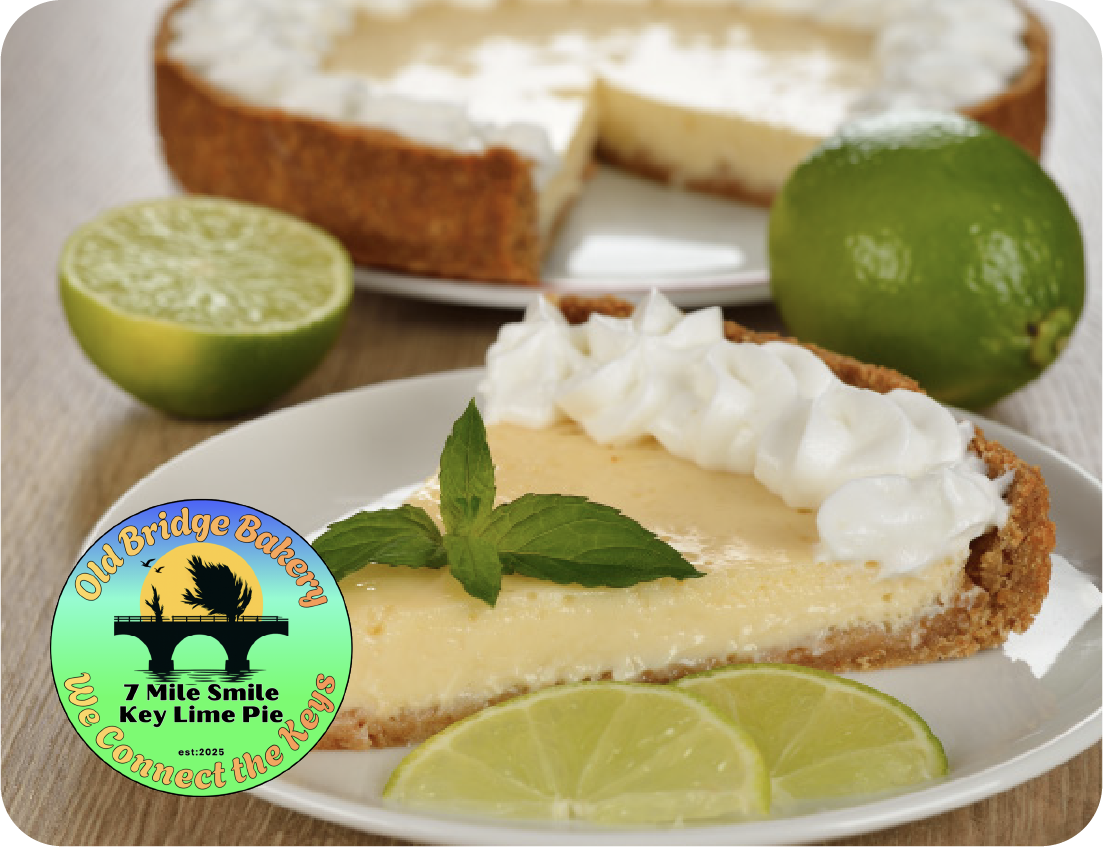 A slice of key lime pie with whipped cream and mint leaves on a plate, garnished with lime wedges. Whole and halved limes set a fresh scene, perfect after enjoying Old Bridge Bakery’s Seafood Showcase. Logo appears in the lower left corner.