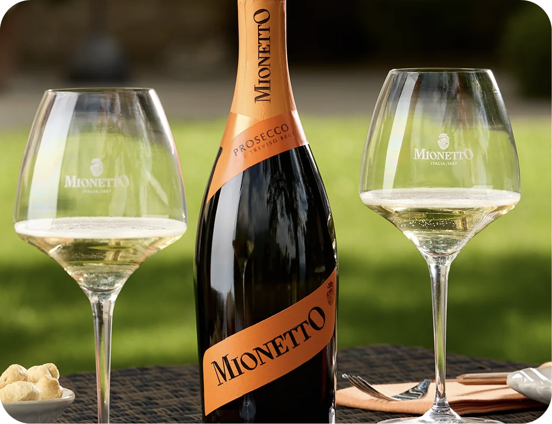 A bottle of Mionetto Prosecco stands between two filled wine glasses on an outdoor table, accompanied by a fork and small seafood display. The background is blurred and grassy.