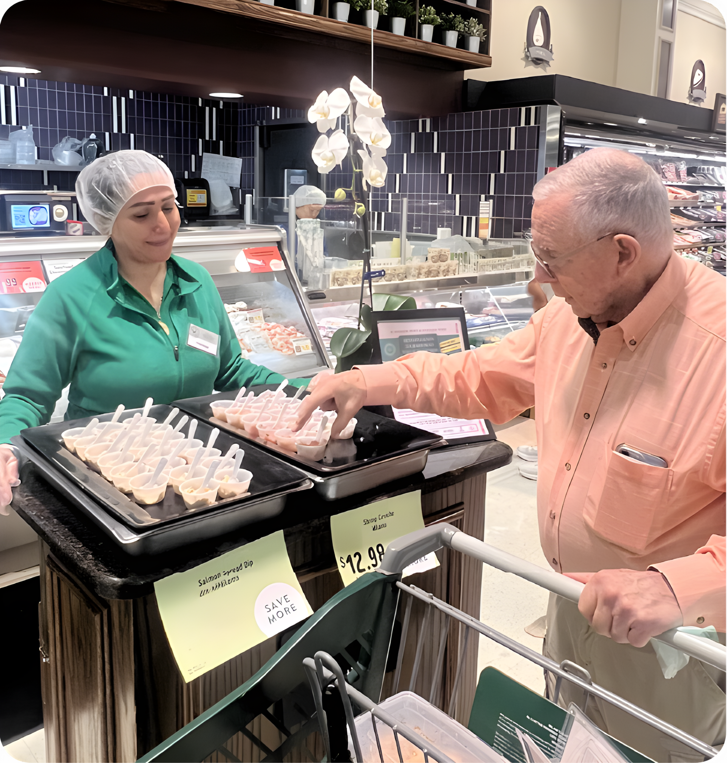 A woman in a green shirt and hairnet offers seafood samples on trays to an older man in an orange shirt at a grocery store deli counter. The man is picking up a sample with a toothpick during the Seafood Showcase.