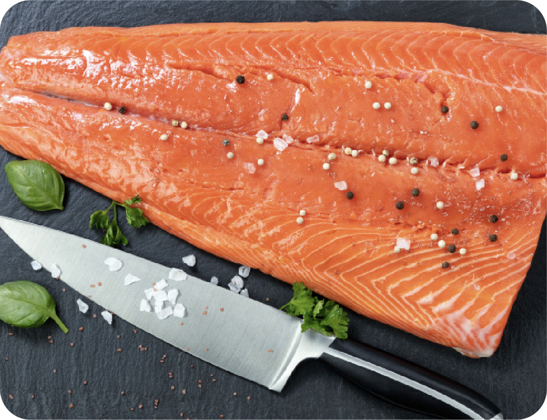 A raw salmon fillet, ready for a Seafood Showcase, is sprinkled with salt and peppercorns on a dark surface beside a chef’s knife, spinach leaves, and fresh herbs.