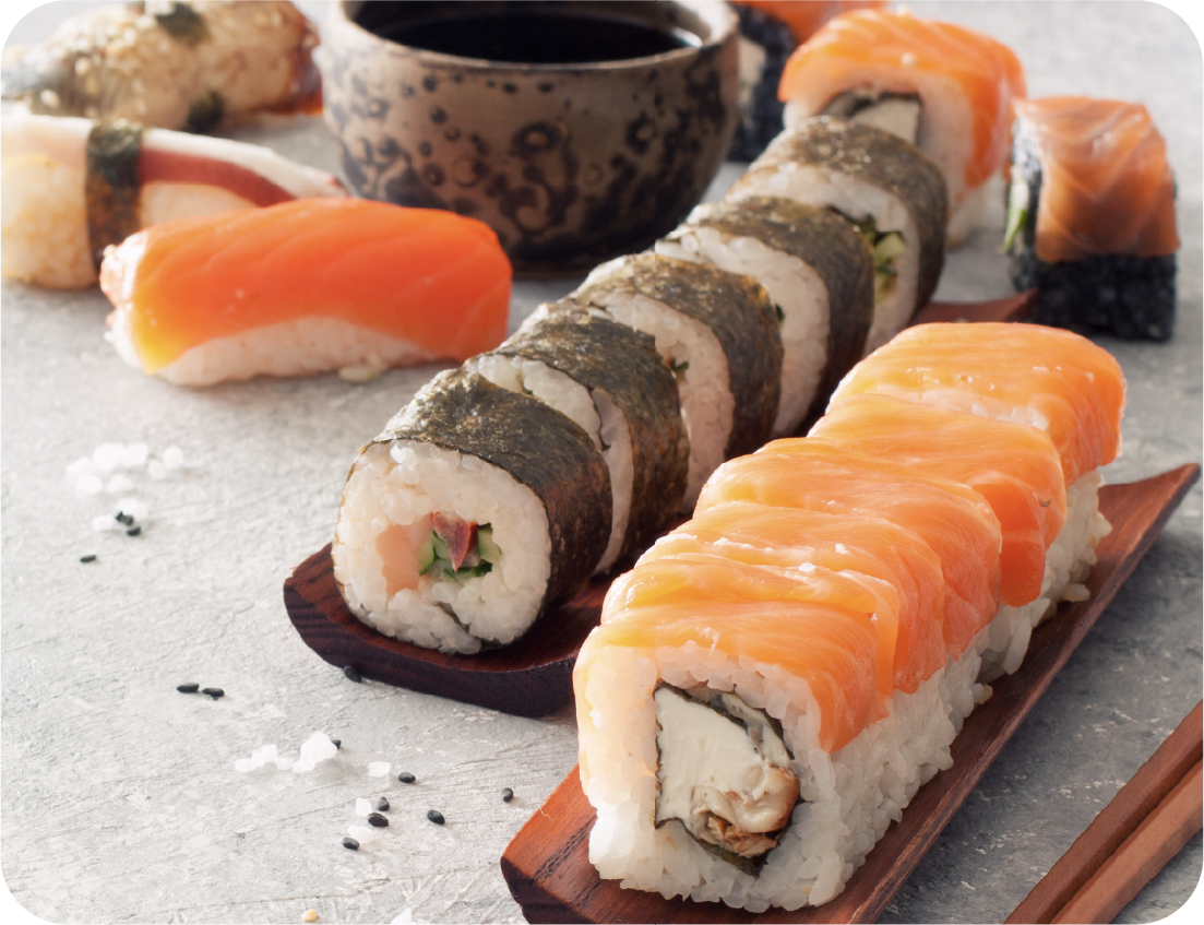 An assortment of sushi rolls and nigiri with salmon, cream cheese, and avocado is arranged on wooden trays beside a bowl of soy sauce, creating a stunning Seafood Showcase with scattered sesame seeds on a light surface.