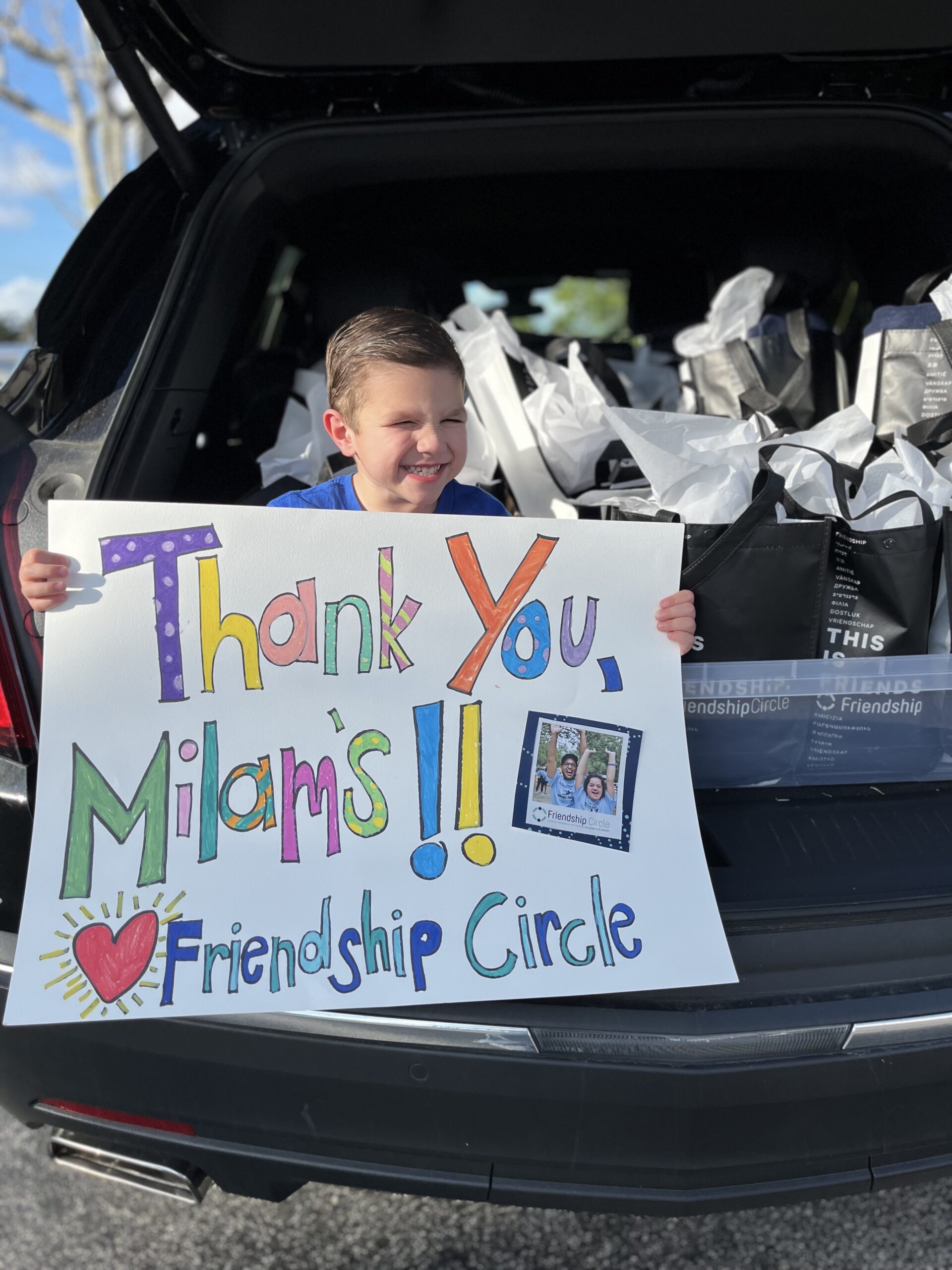Smiling child holds a colorful Thank You, Milams!! ♥ Friendship Circle sign in front of an open car trunk filled with black and white bags, celebrating local donations. A small photo is attached to the sign. Sunny day.