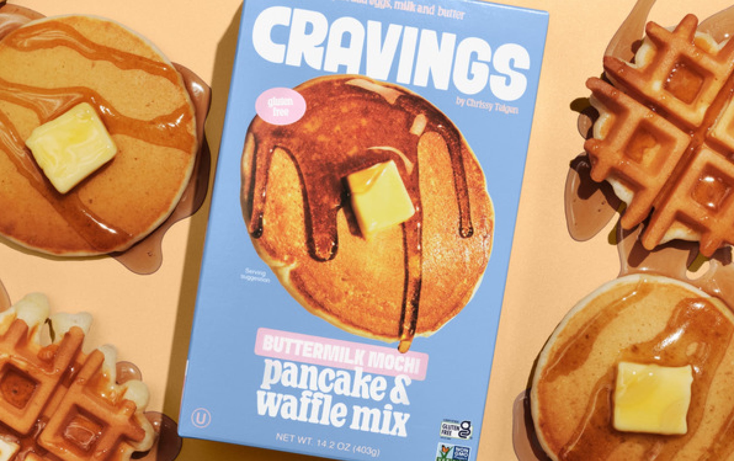 A box of Cravings Buttermilk Mochi Pancake & Waffle Mix is surrounded by pancakes and waffles topped with butter and syrup on a light orange surface.