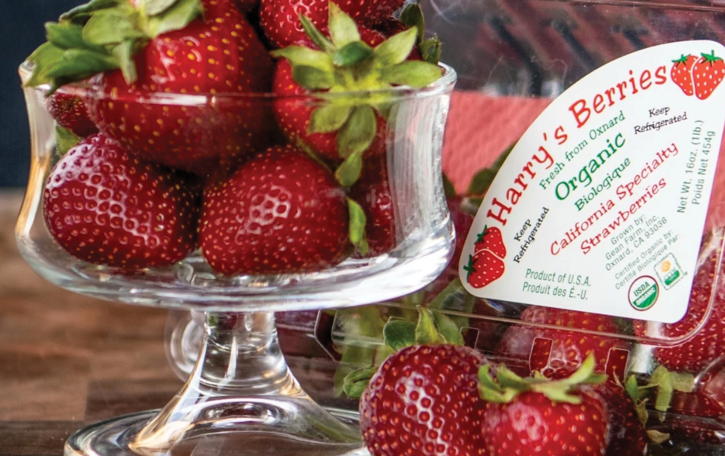 A glass dish filled with fresh strawberries sits beside a plastic container labeled Harrys Berries Organic California Specialty Strawberries. Some strawberries are also scattered on the wooden surface.