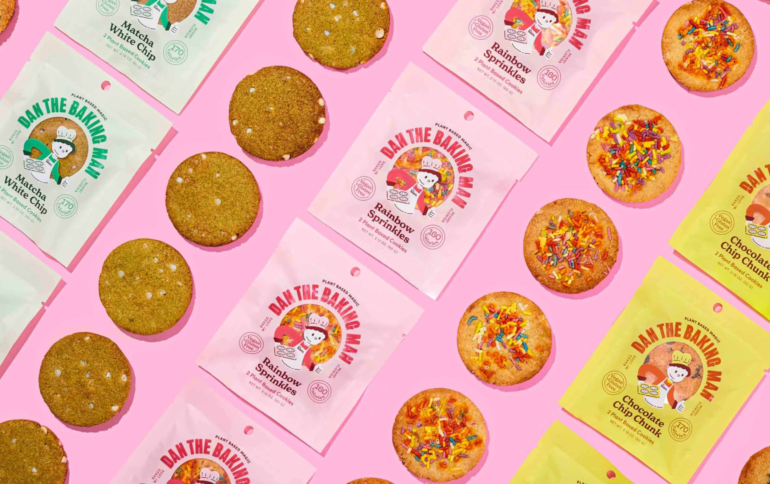Flat lay of cookies and colorful cookie packages on a pink background. Packages are yellow and pink, labeled “Dan the Baking Man,” with illustrated faces and flavors like Birthday Sprinkle and Matcha White Chip.