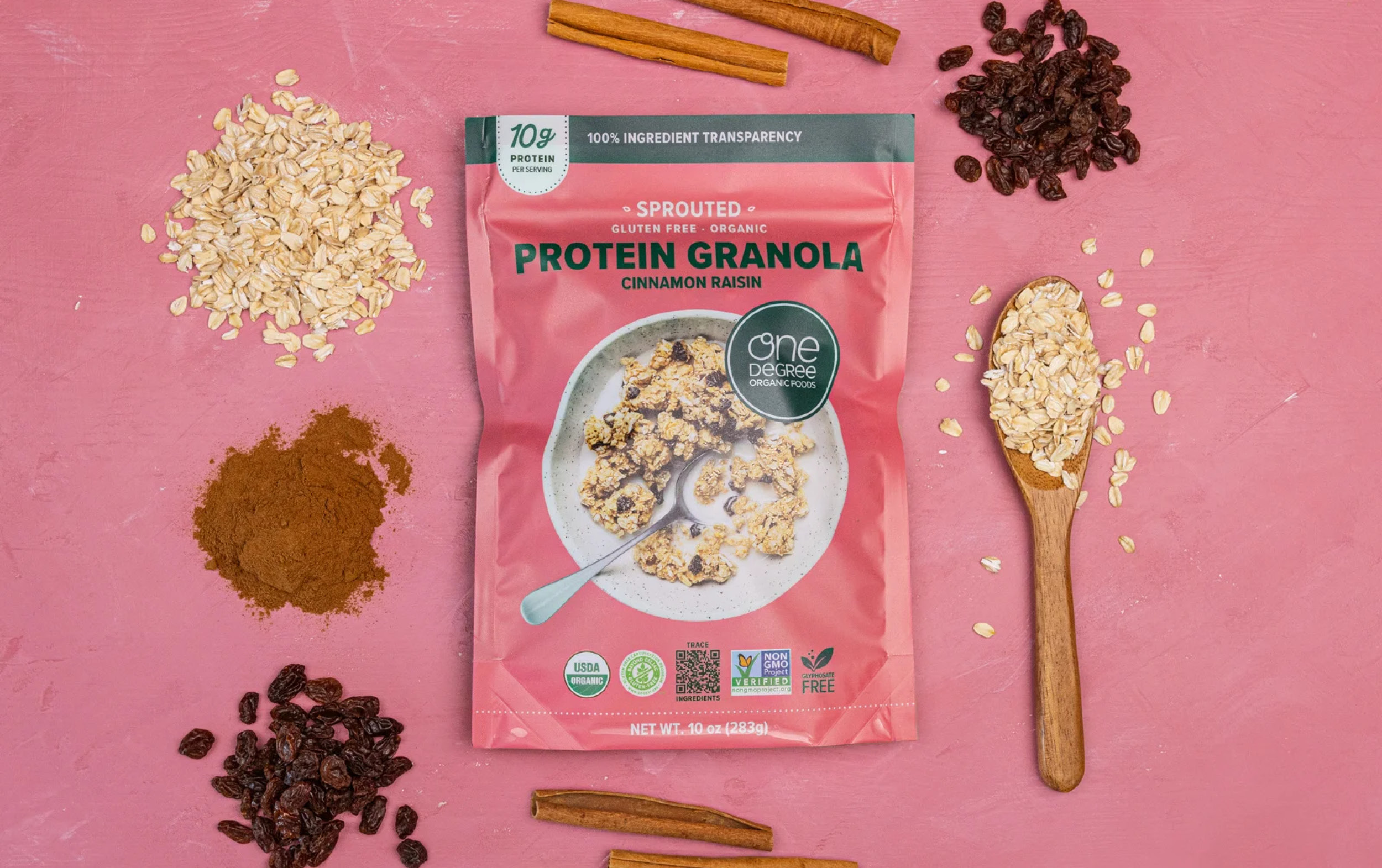 A pink package of One Degree Organic Foods Cinnamon Raisin Protein Granola is on a pink surface, surrounded by oats, raisins, cinnamon sticks, a heap of ground cinnamon, and a wooden spoon with oats.