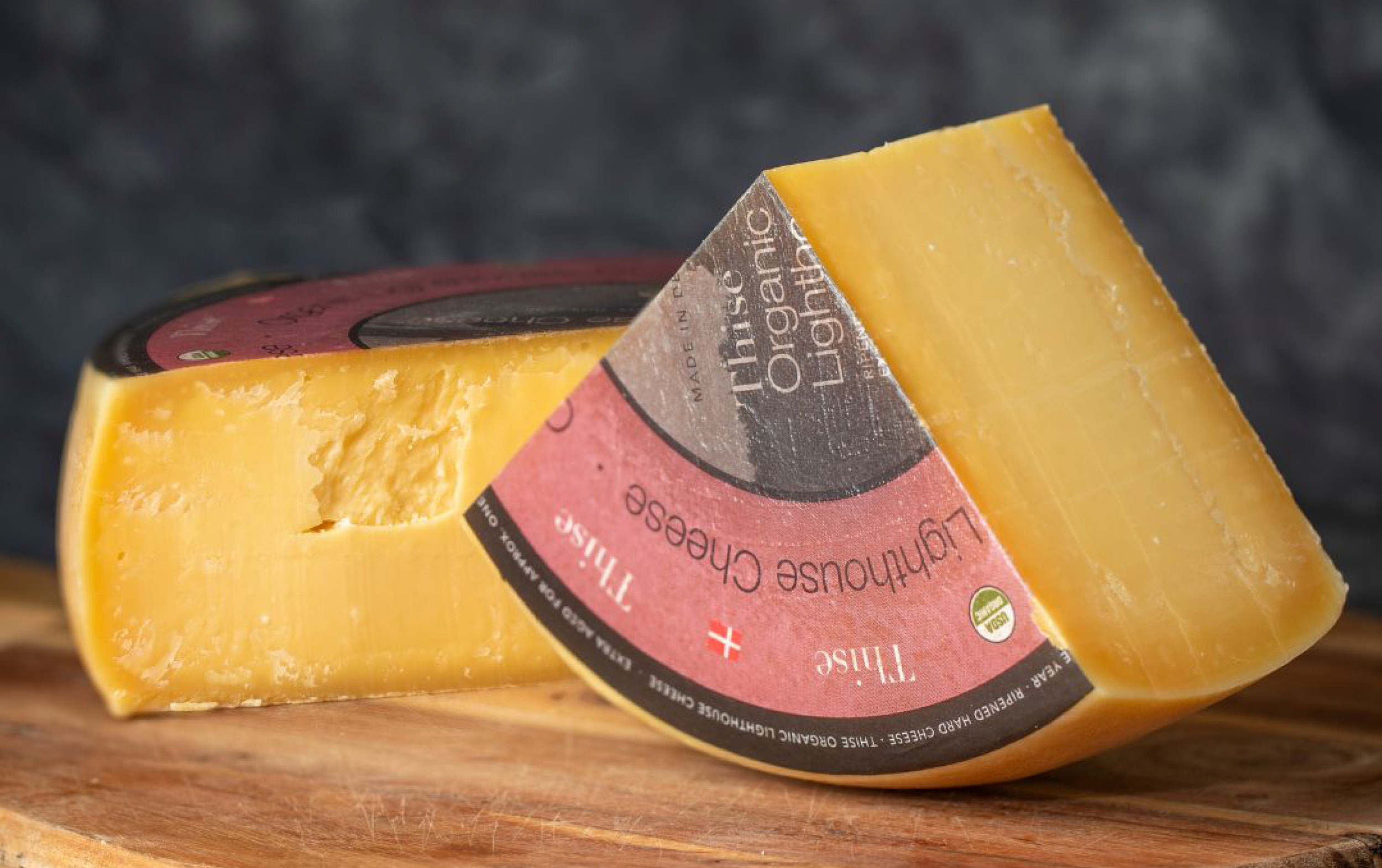 A large wheel of yellow cheese, partially cut, with a brown and red label on the rind, sits on a wooden board against a dark, blurred background.
