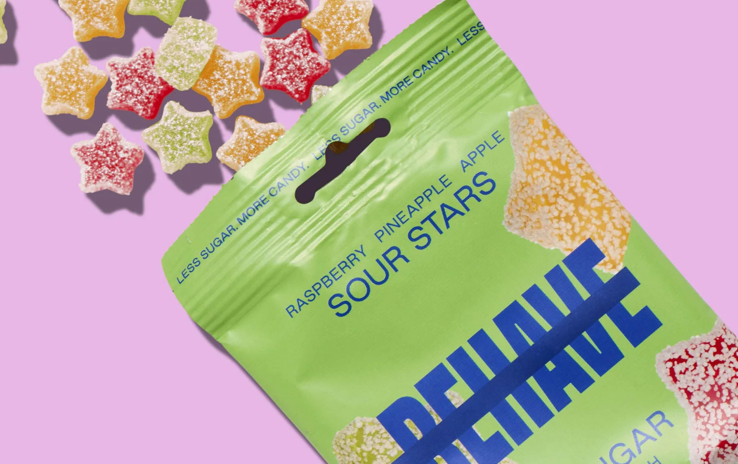 A green bag labeled BEHAVE Sour Stars with colorful, sugar-coated star-shaped gummies spilling out onto a pink background.