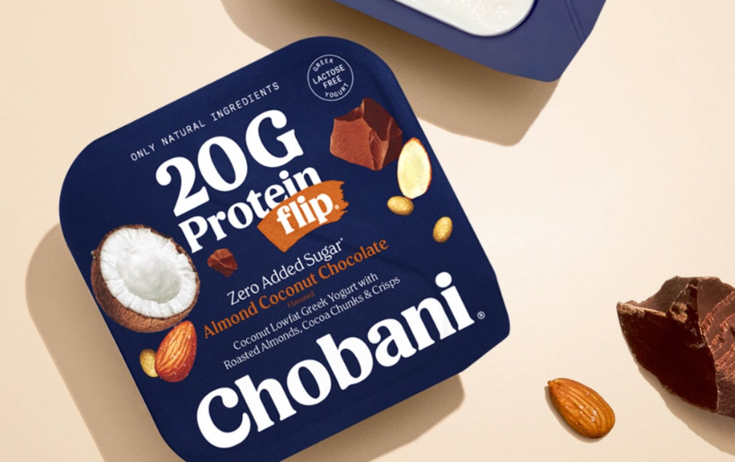 Chobani 20G Protein Flip yogurt, Almond Coconut Chocolate flavor, with images of coconut, chocolate, and almonds on dark blue packaging. Yogurt cup and ingredients shown on a light beige surface.
