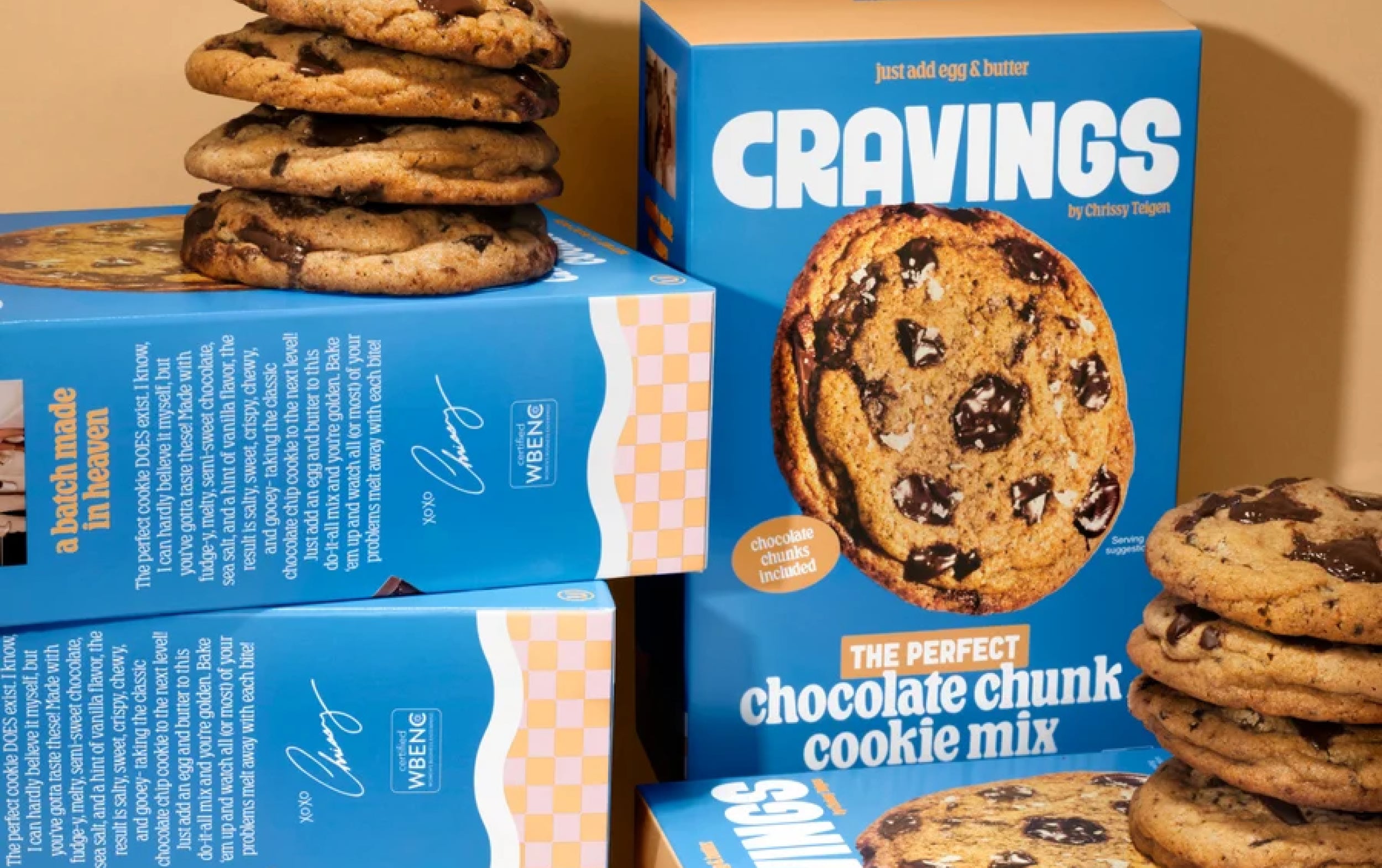 Boxes of Cravings chocolate chunk cookie mix are surrounded by stacks of chocolate chip cookies, with one cookie leaning against a box. The packaging is blue with a cookie image and text highlighting the product features.