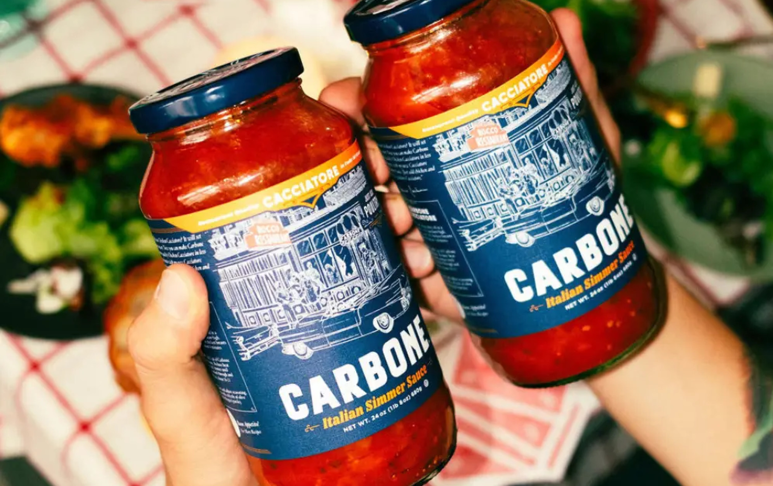 Two hands hold jars of Carbone Italian simmer sauce with blue and yellow labels, set against a blurred background with food and a red-and-white checkered tablecloth.
