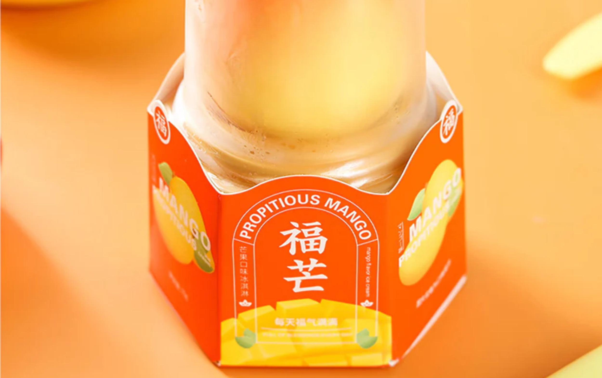 A glass bottle with a bright orange hexagonal label reading Propitious Mango and Chinese characters, against an orange background. The label features images of mangoes and yellow accents.