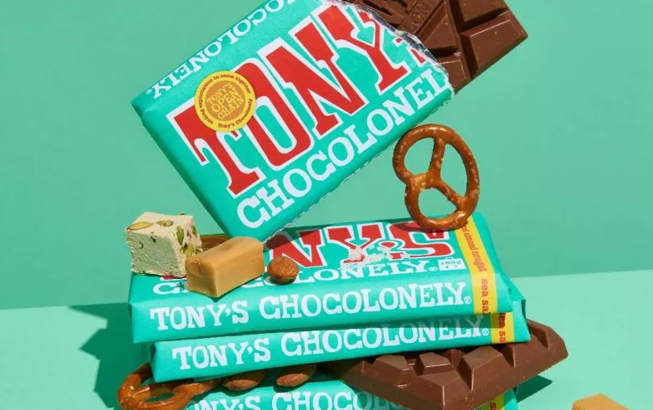 Three stacked Tony’s Chocolonely chocolate bars with turquoise wrappers are surrounded by pieces of chocolate, fudge, nougat, and pretzels, set against a green background.