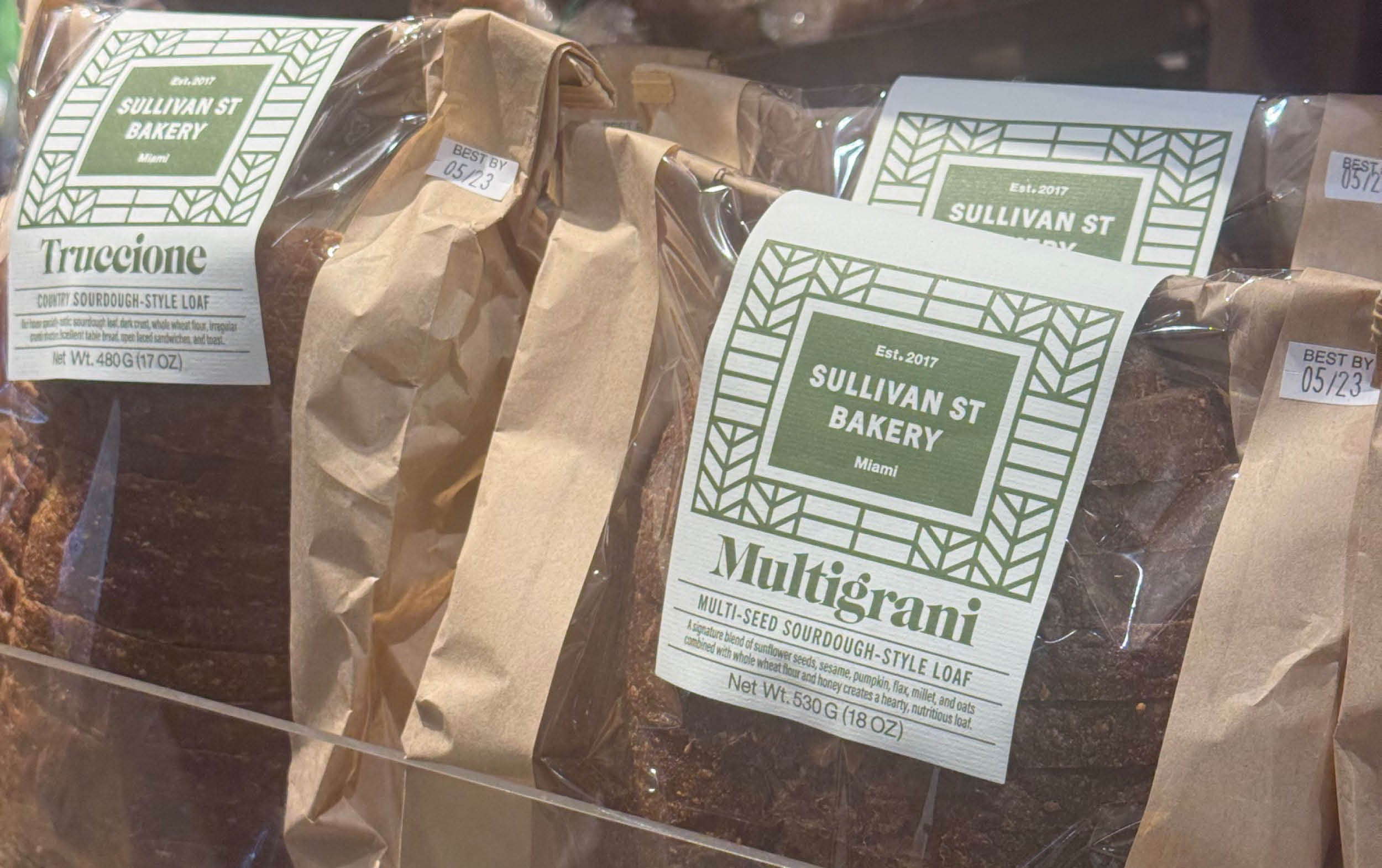 Several loaves of bread from Sullivan St Bakery, including Truccione and Multigrani varieties, are displayed in paper bags with green and white labels in a bakery case.