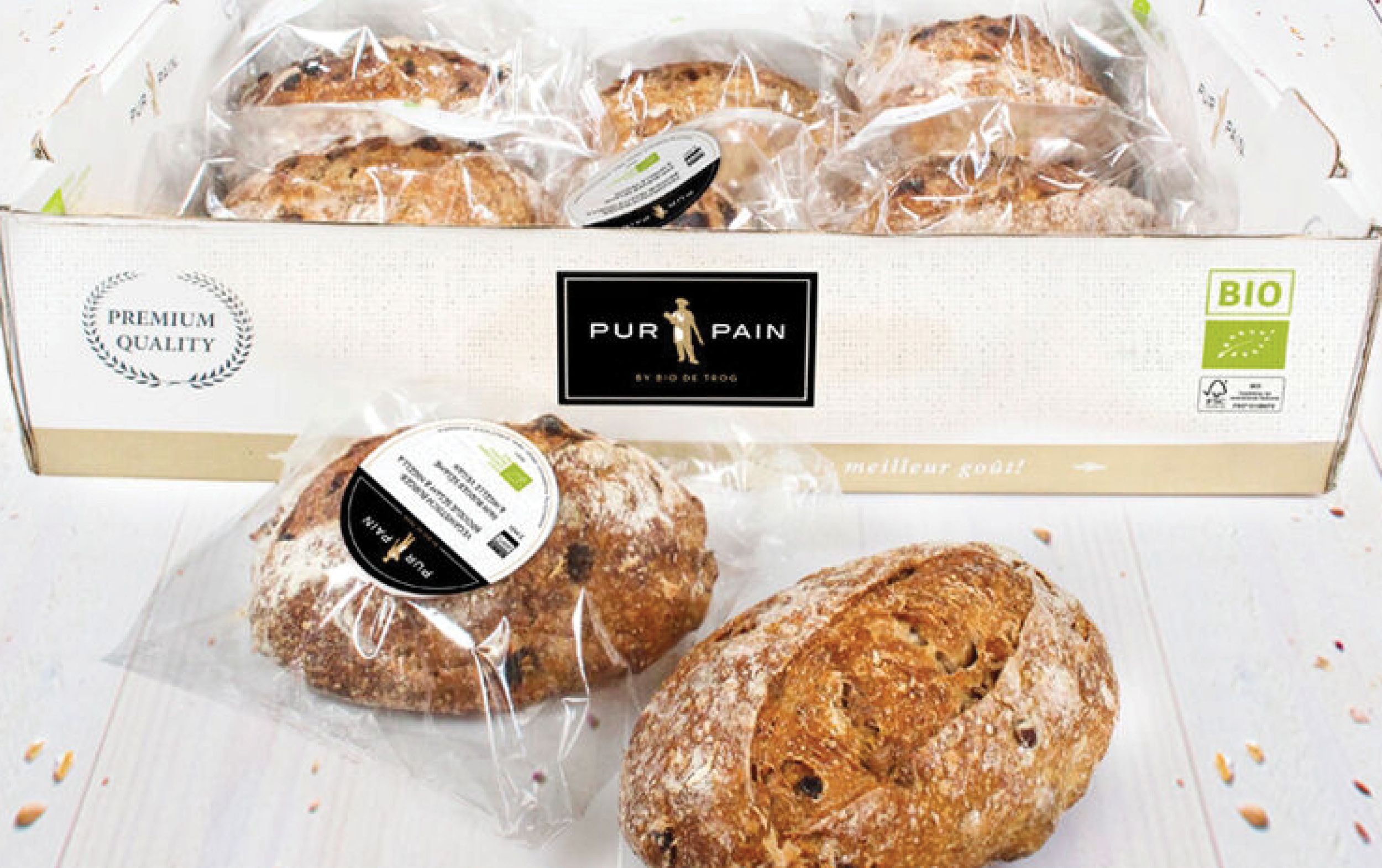 Six loaves of artisanal bread, individually wrapped in clear plastic, are displayed in a white box labeled “PUR PAIN.” Two loaves are placed outside the box. The packaging features “Premium Quality” and organic certification labels.