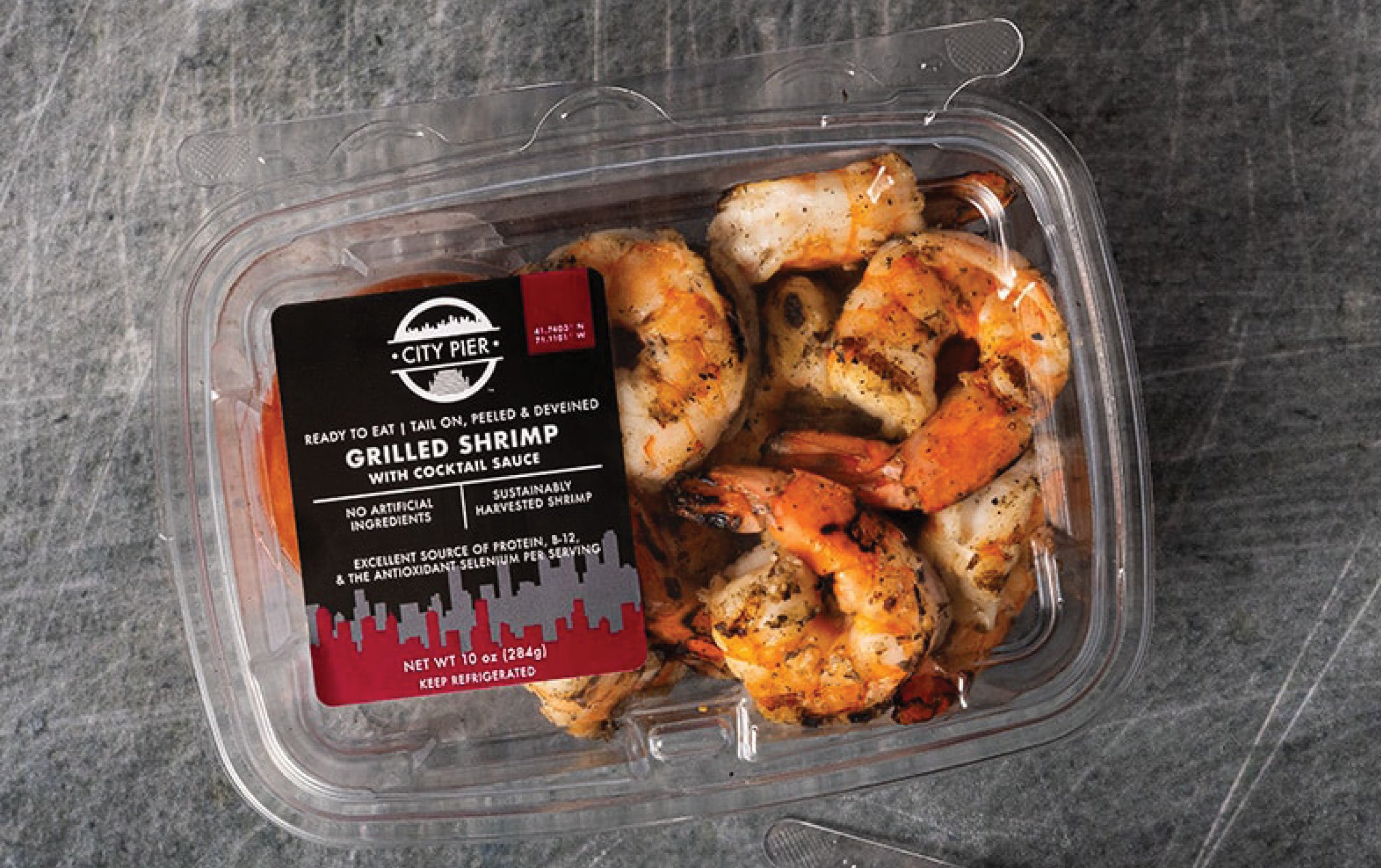 A clear plastic container of City Pier grilled shrimp with a black and red label, showing cooked shrimp inside, placed on a dark gray textured surface.