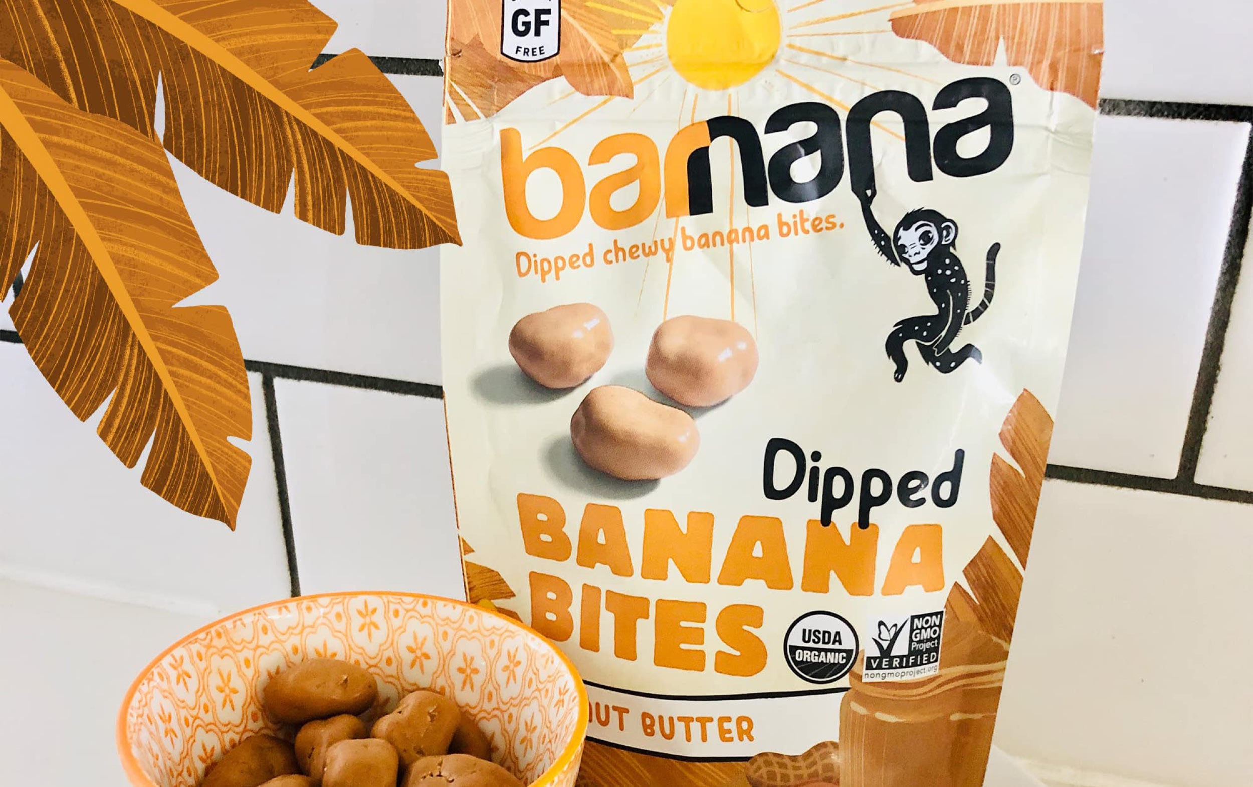 A bag labeled banana dipped banana bites with images of coated bites, a monkey, and certification logos. An orange-patterned bowl filled with the treats sits in front. Brown palm leaves are partly visible on the edge.