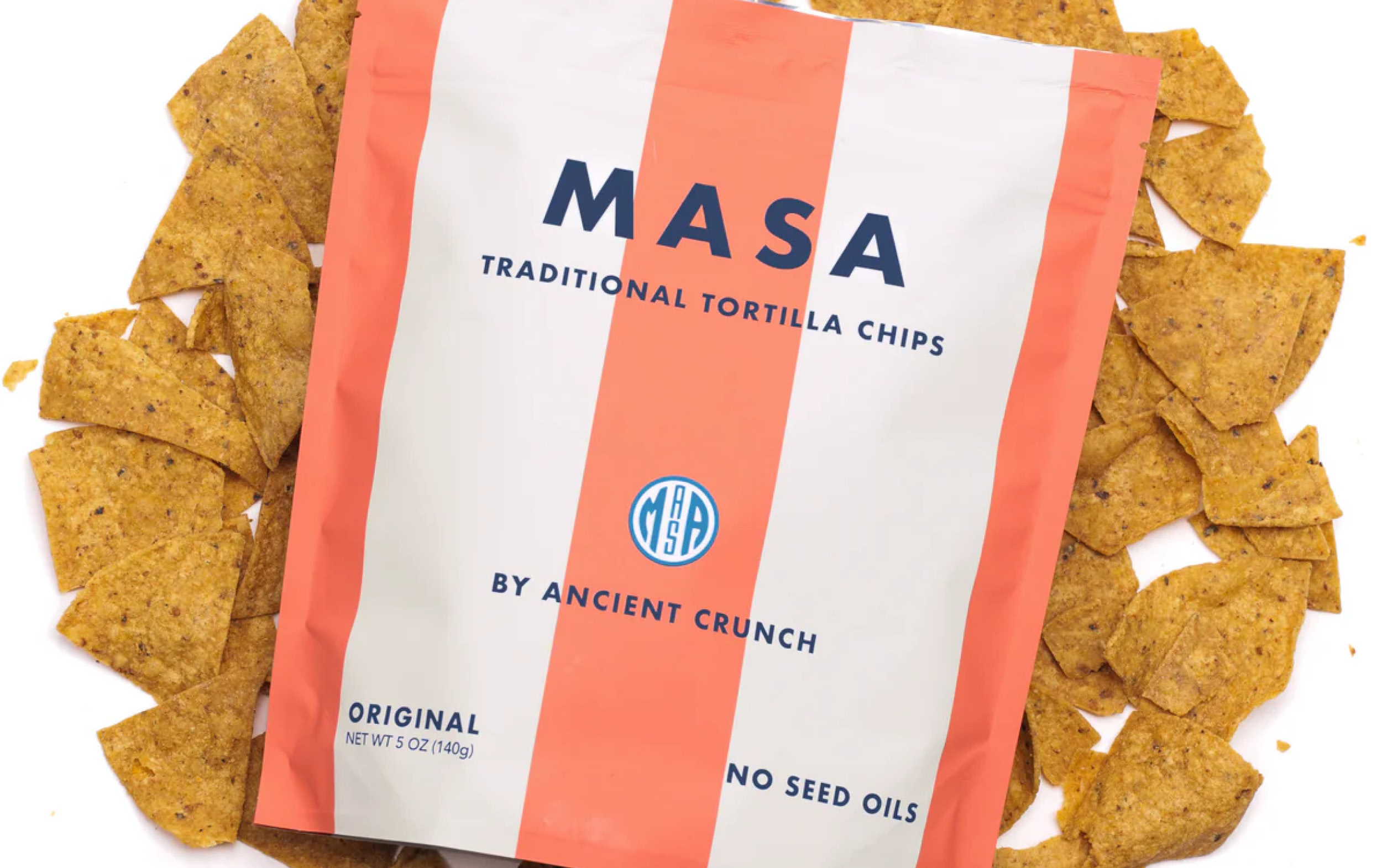 A bag of MASA traditional tortilla chips by Ancient Crunch sits on a pile of chips. The bag is cream and orange with blue text, stating “No Seed Oils” and “Original.”.