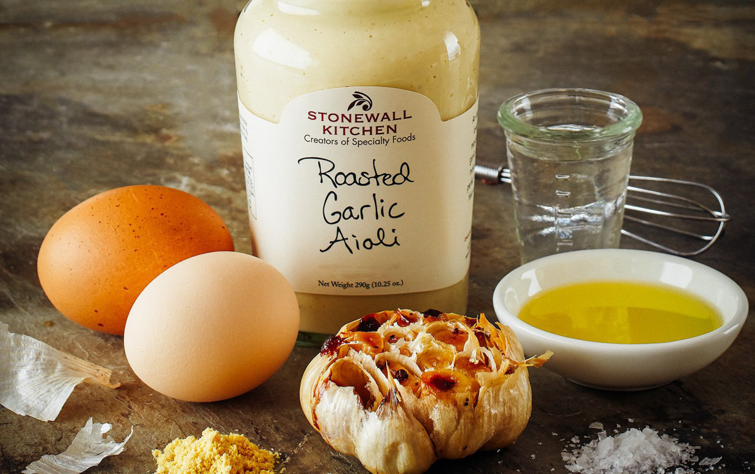 A jar labeled Roasted Garlic Aioli is surrounded by two eggs, roasted garlic, a small glass of olive oil, salt, mustard powder, and a metal whisk on a wooden surface.