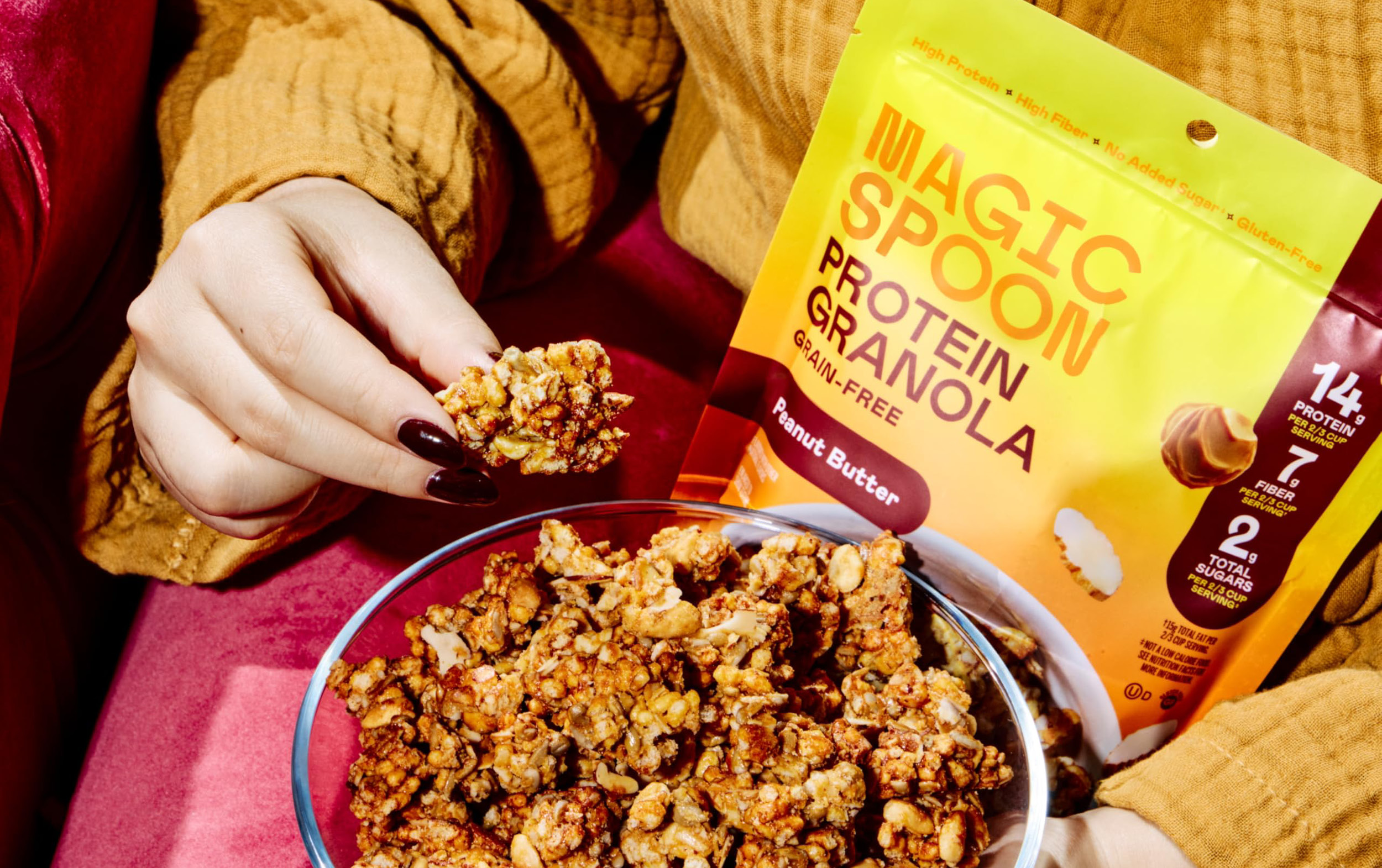 A person holding a cluster of peanut butter protein granola above a bowl, with a bag of Magic Spoon Protein Granola in the background. The person is wearing a mustard-colored garment.