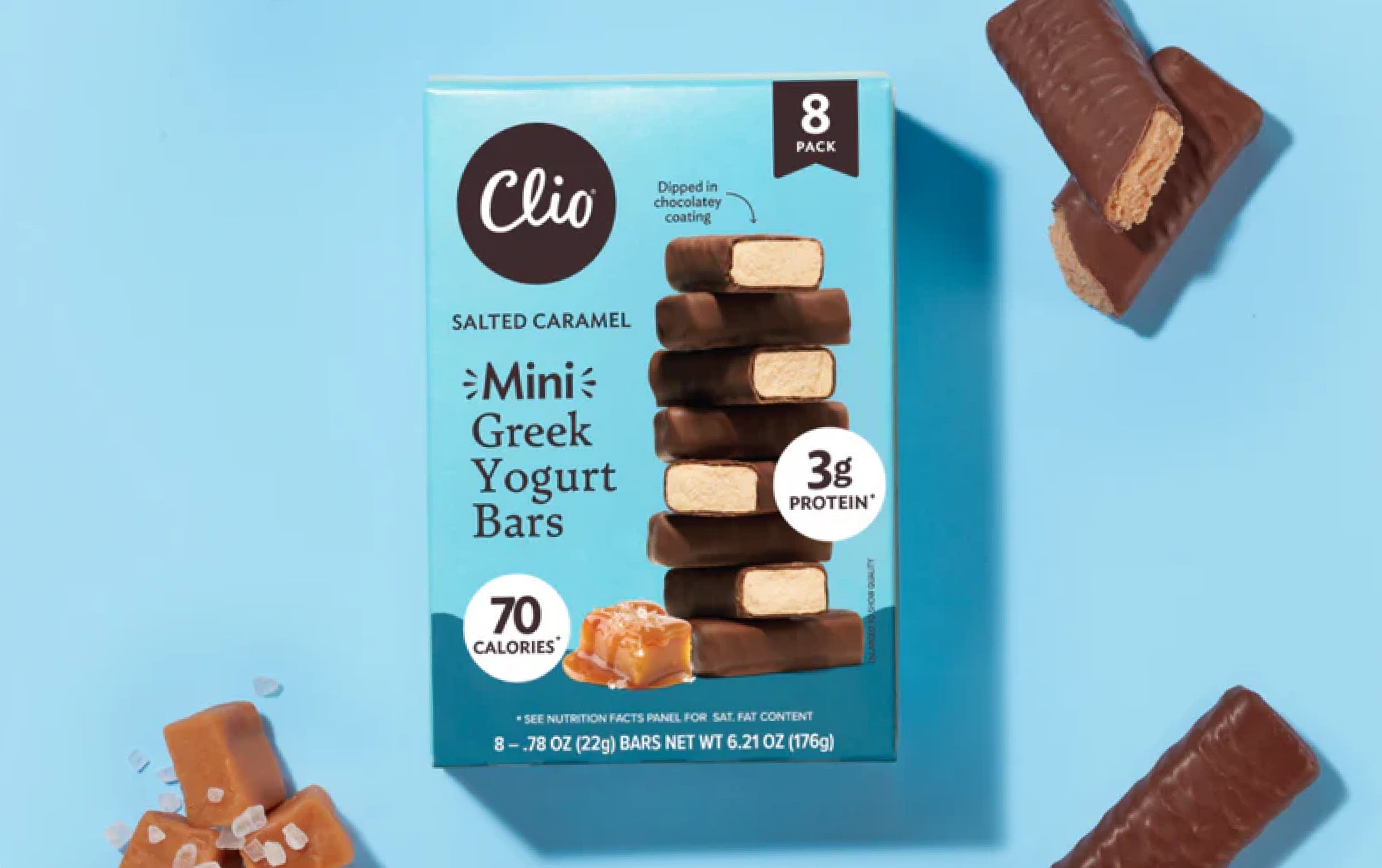 A box of Clio Salted Caramel Mini Greek Yogurt Bars sits on a blue surface, surrounded by chocolate-covered bars and caramel pieces. The box highlights 3g protein, 70 calories, and contains eight bars.