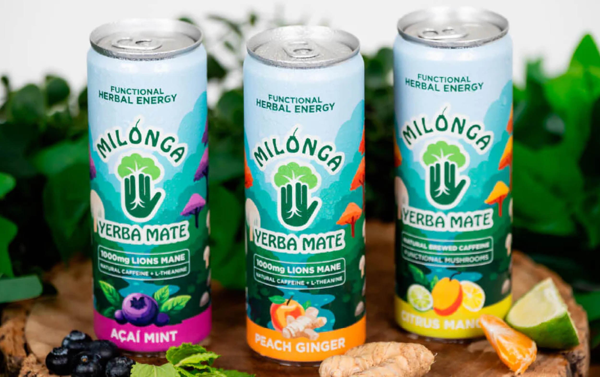 Three cans of Milonga Yerba Mate drinks in acai mint, peach ginger, and citrus mango flavors are displayed on a wooden surface with fresh fruits, ginger, and greens around them.