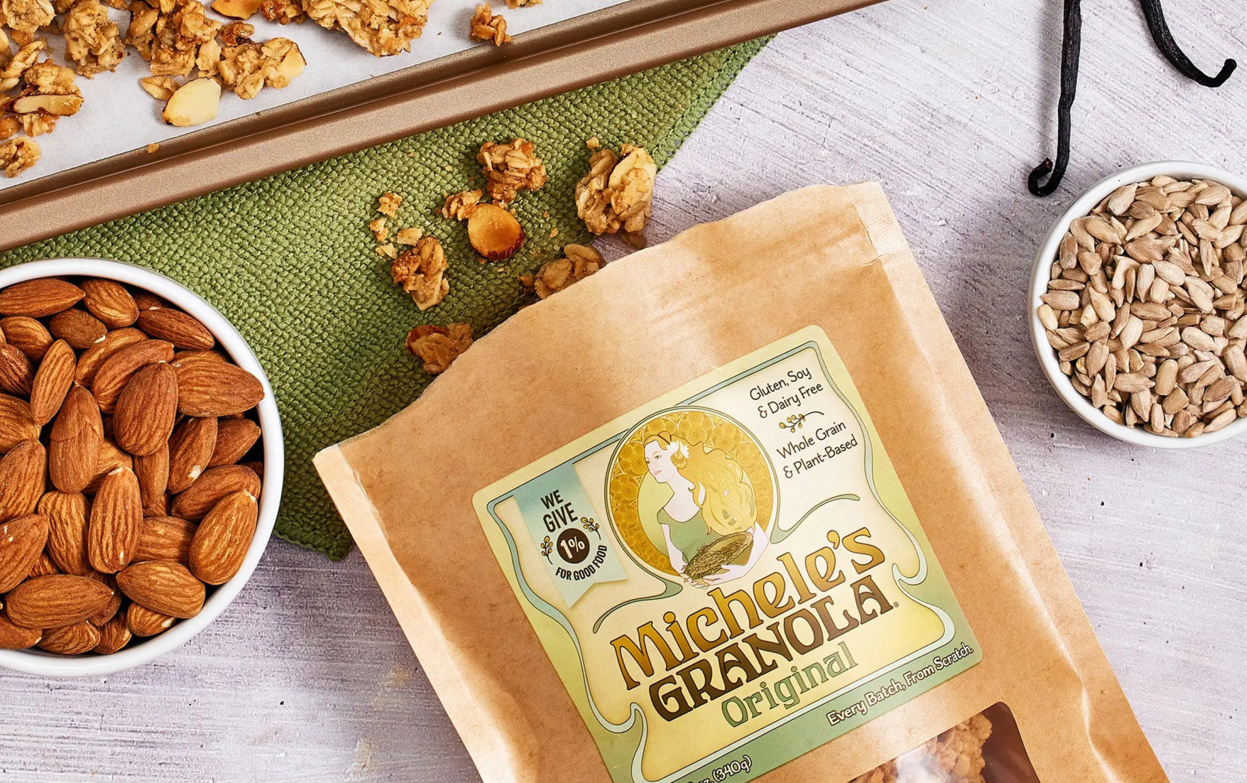 A bag of Michele’s Granola Original flavor sits on a table with almonds, sunflower seeds in bowls, and a baking tray with granola pieces on a green cloth nearby.