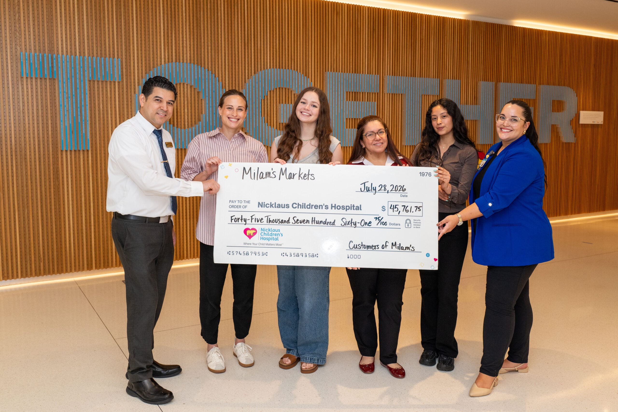 Six people stand indoors holding a large donation check for $45,761.61 made to Nicklaus Children’s Hospital. This wonderful act exemplifies the power of Community Donations, with the word TOGETHER appearing in large letters on the wall behind them.