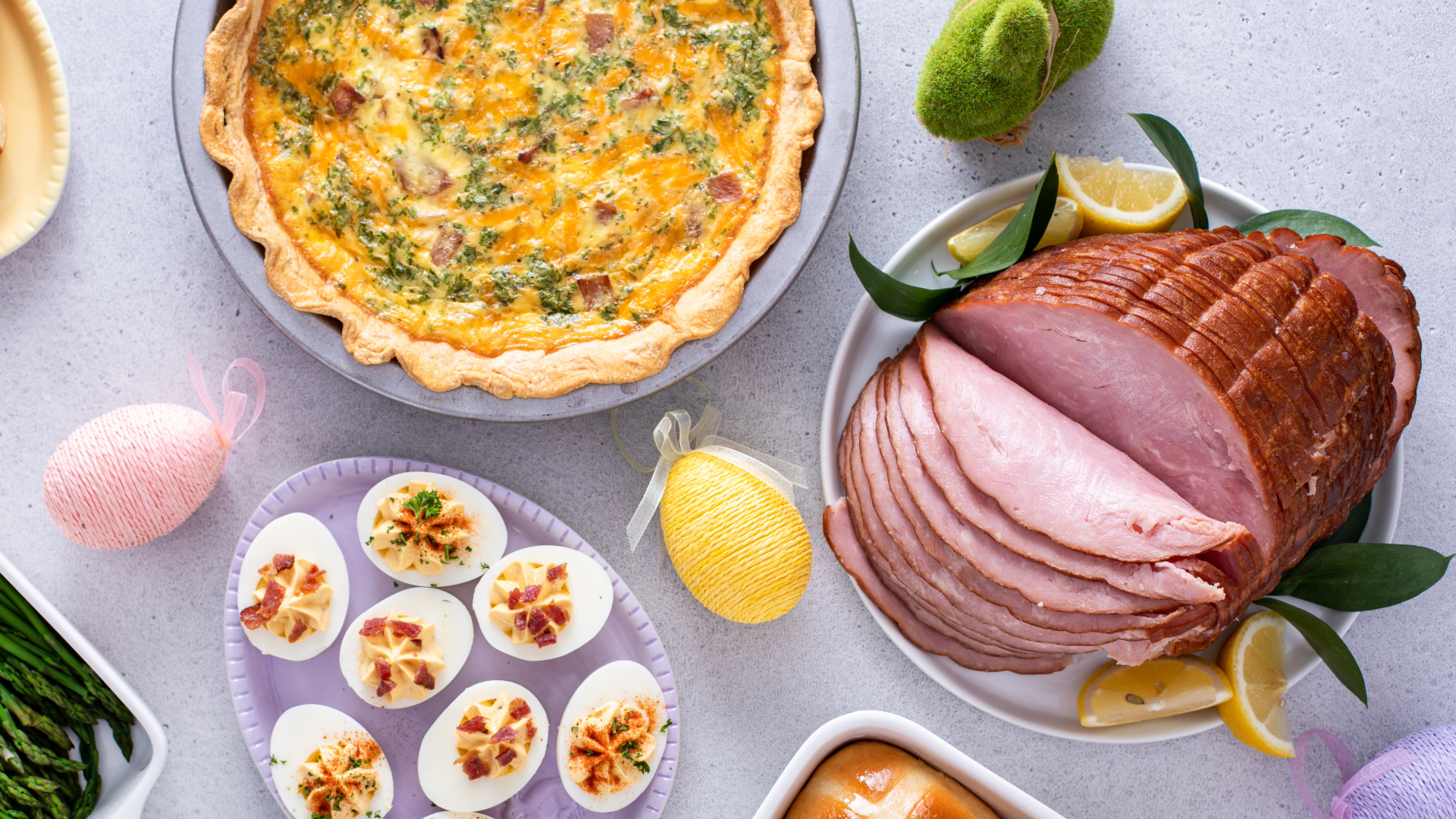 A table set with a quiche, sliced glazed ham with lemon wedges, deviled eggs on a tray, asparagus, and decorative Easter eggs creates a festive spring meal you can now order online for easy holiday entertaining.