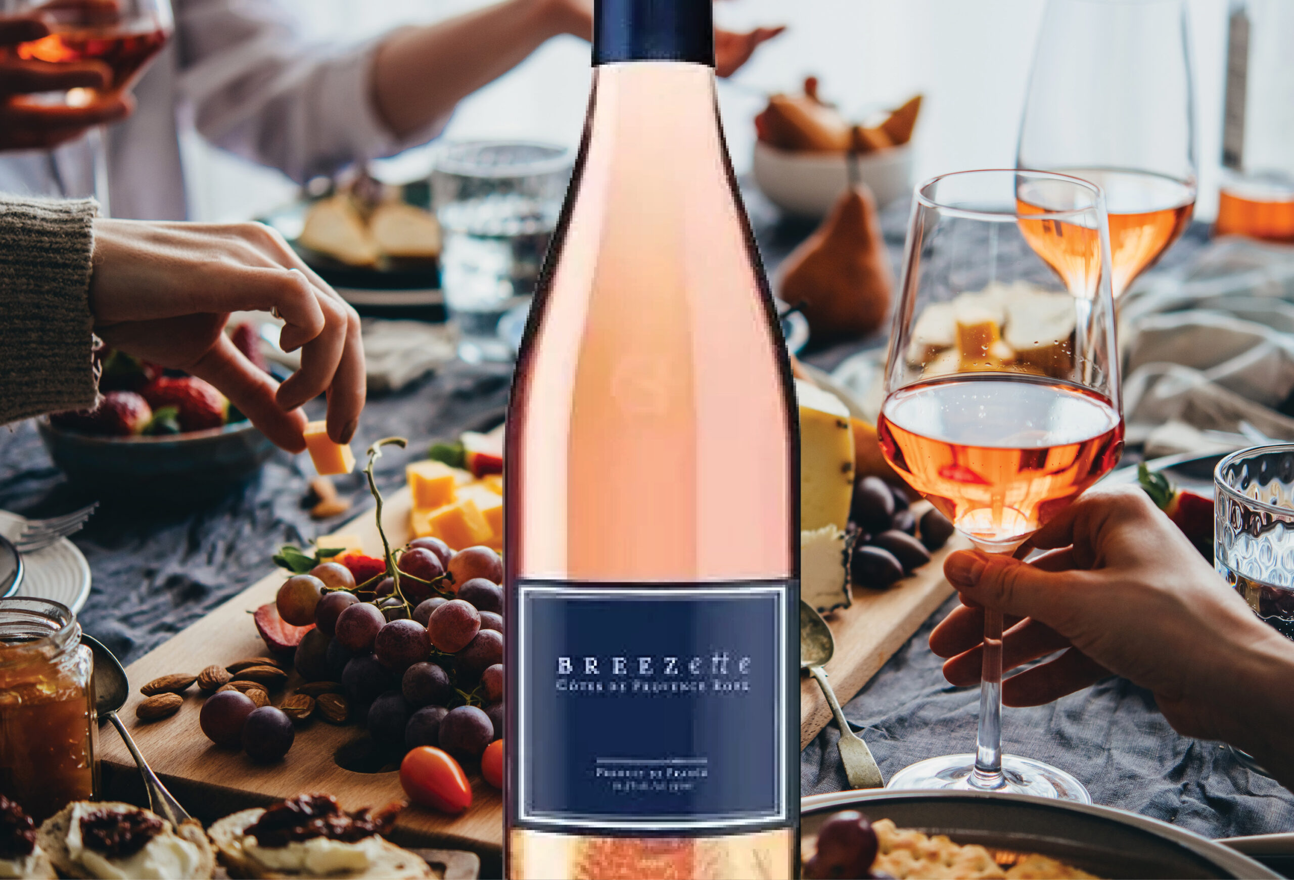 A bottle of BREEzette rosé wine on a table with snacks, fruits, and cheese, surrounded by people holding glasses of rosé, sharing a meal together.