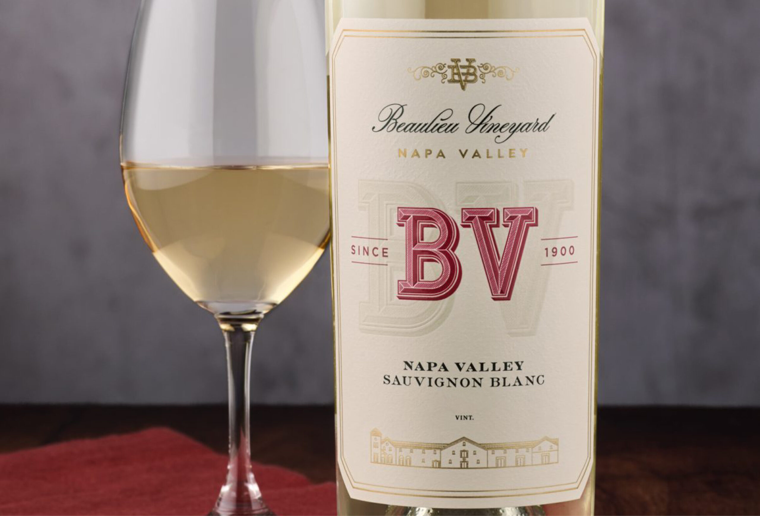 A glass of white wine sits next to a bottle labeled Beaulieu Vineyard Napa Valley Sauvignon Blanc with a red and gold label and the initials BV, perfect for those seeking exclusive Wine Deals or Buy One Get One Wines offers.