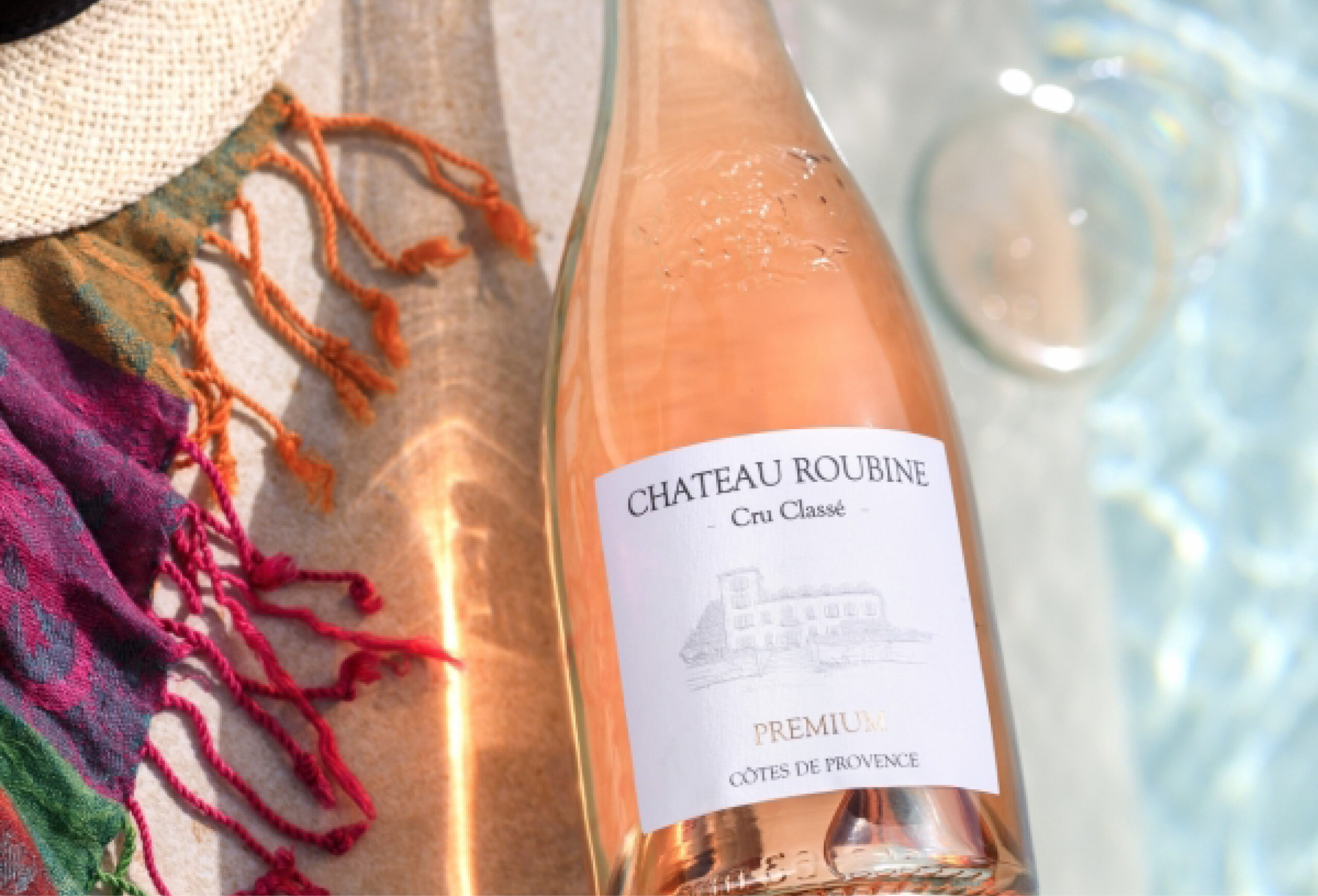 A bottle of Château Roubine rosé wine lies by a poolside, next to a colorful scarf and a straw hat, with sunlight reflecting on the water—perfect for enjoying exclusive Wine Deals with a glass in the background.