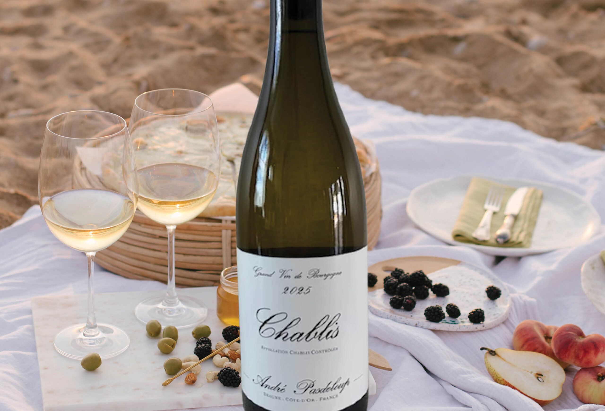 A bottle of Chablis wine stands on a beach picnic setup with two filled wine glasses, cheese, olives, blackberries, and sliced fruit on a white cloth over the sand. Plates and cutlery are nearby.