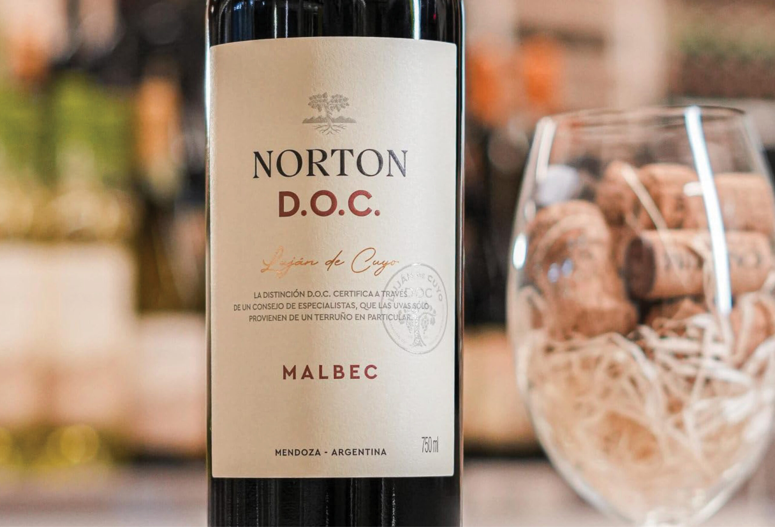 A bottle of Norton D.O.C. Malbec wine from Mendoza, Argentina is displayed next to a wine glass filled with corks. The background is softly blurred with warm tones—perfect for those looking to discover quality wines or enjoy a Buy One Get One Free offer.