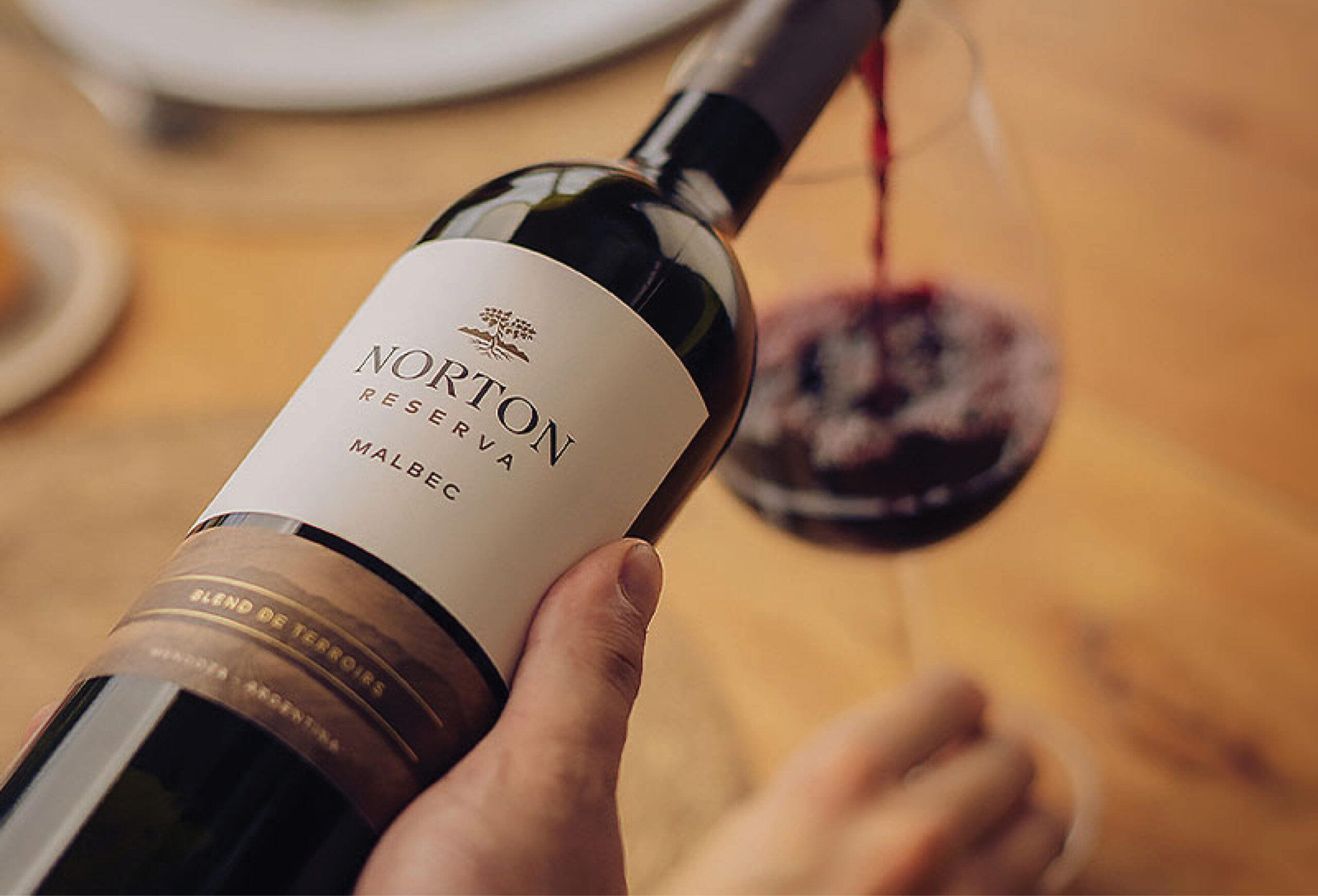 A hand holds a bottle of Norton Reserva Malbec wine as red wine is poured into a glass on a wooden table, showcasing an irresistible Buy One Get One offer for wine lovers.