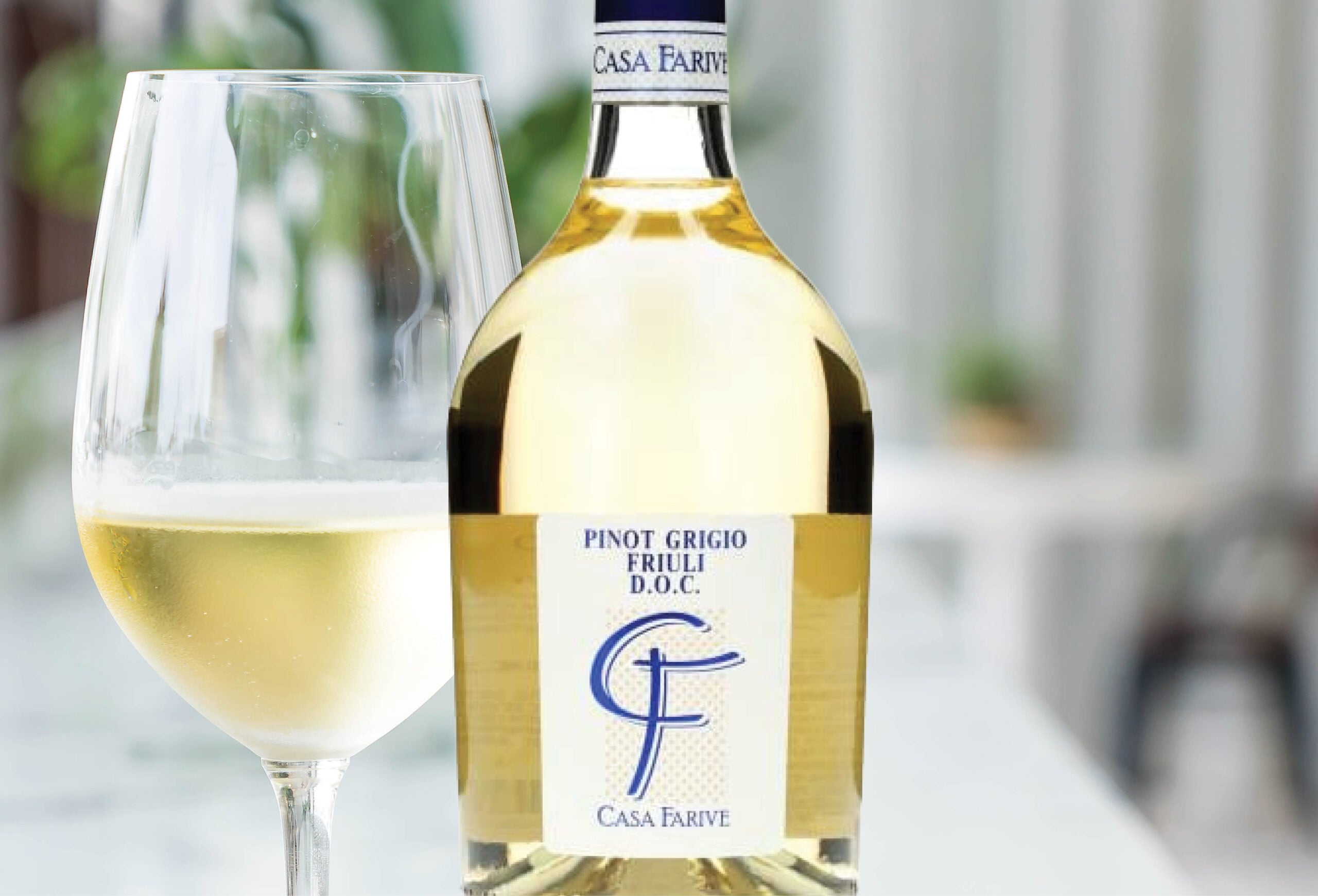 A glass of white wine sits next to a bottle labeled Pinot Grigio Friuli D.O.C. Casa Farive on a blurred outdoor table, inviting you to savor refreshing flavors—perfect for those seeking fantastic wine deals or buy one get one free wines.