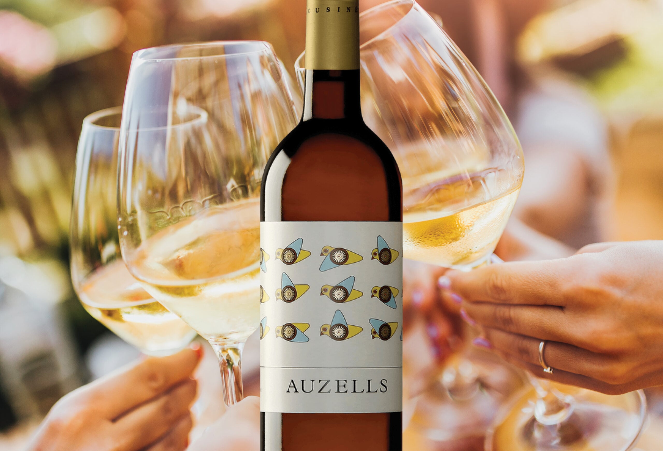 A bottle of Auzells white wine is in focus, its label adorned with birds. In the blurred background, three hands clink glasses in a celebratory toast—perfect for enjoying current wine deals.